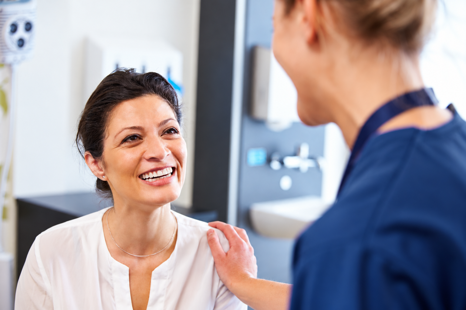 A woman in a white shirt smiling and talking to a healthcare professional in a blue uniform, in a medical setting.