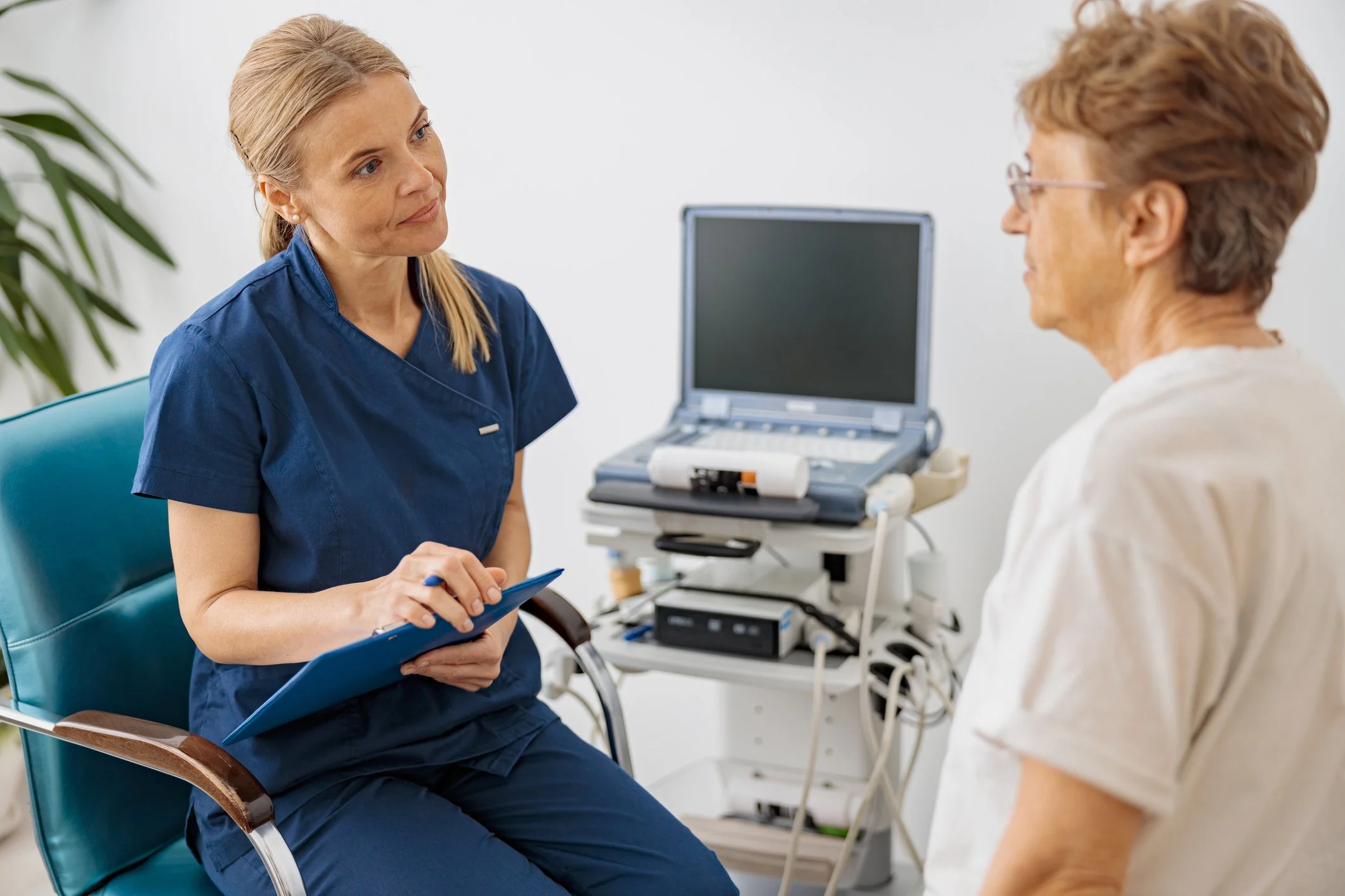 A female healthcare professional in blue scrubs sitting in a chair, holding a clipboard and pen, engaging in conversation with an older patient with short hair and glasses, in a medical examination room with a computer and medical equipment.