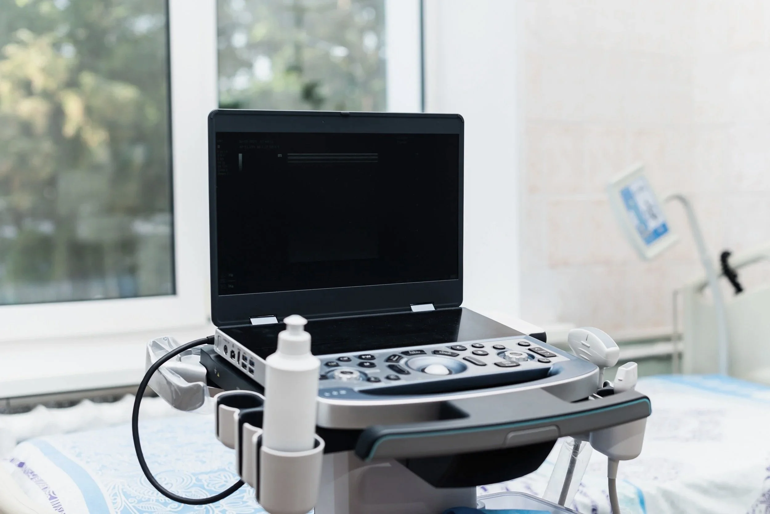Medical ultrasound machine with monitor, probes, and bottles in a clinical setting near a window.