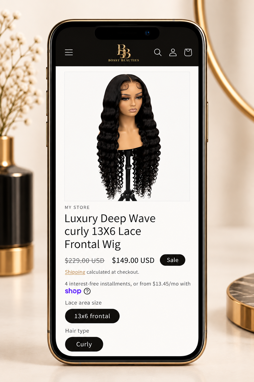 Smartphone displaying an online store page for a luxury deep wave curly wig with lace front, priced at $149, on a white surface with a gold-rimmed mirror and a flower arrangement in the background.