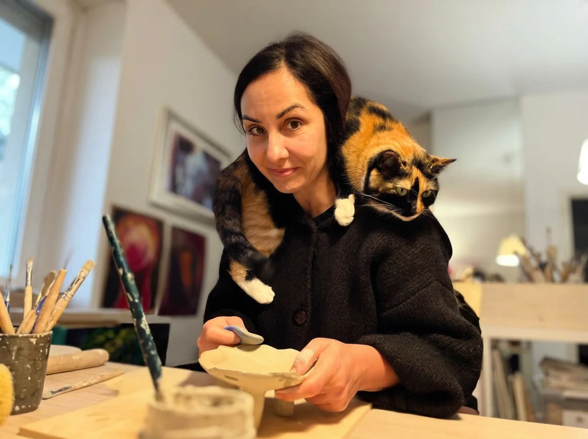 A woman in a black sweater with a calico cat draped over her shoulders, on a wooden table in an art studio or workspace, with brushes and art supplies nearby, and framed artwork on the wall in the background.
