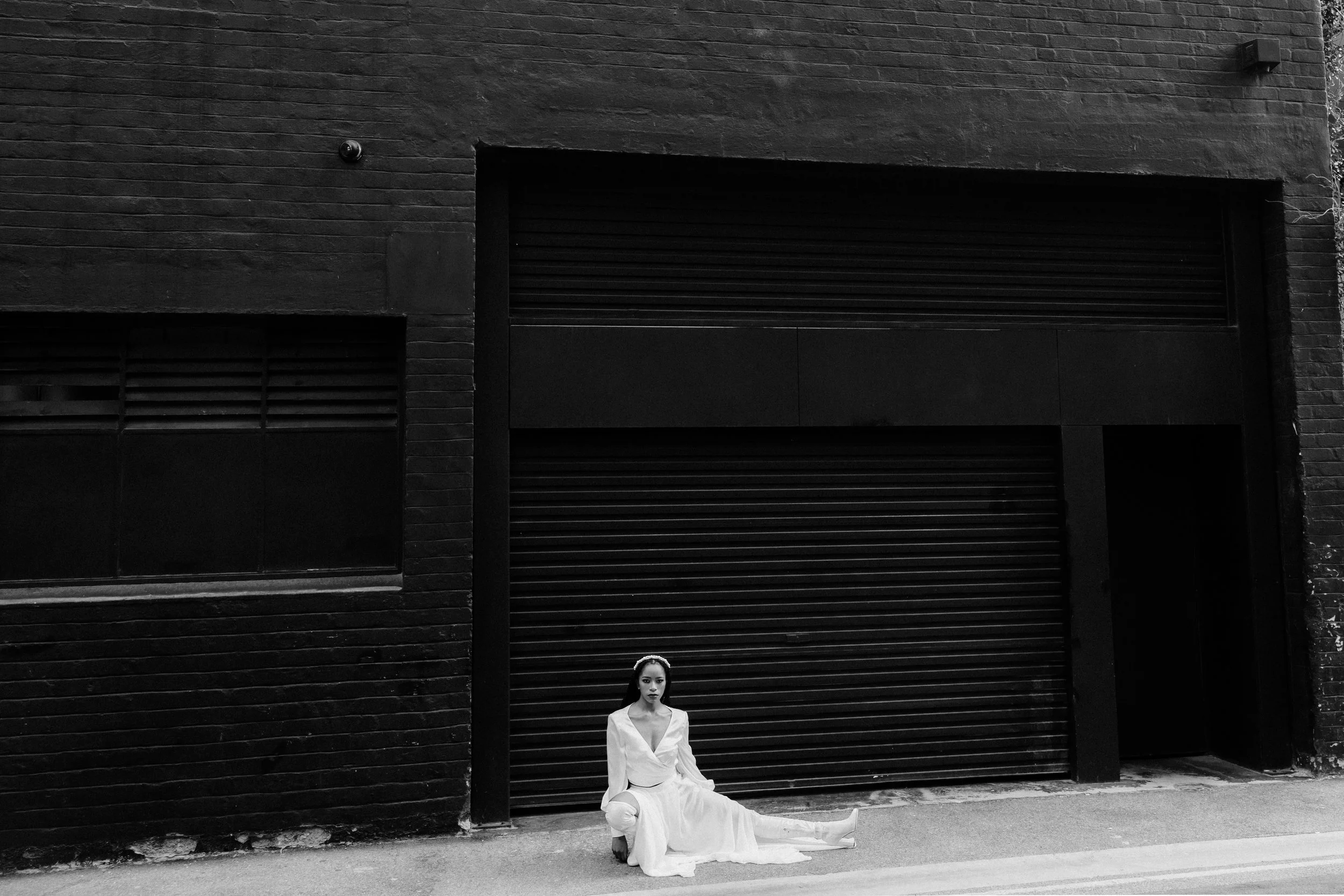 A woman in a white dress sitting against a large black garage door on a city street, with a brick wall and small security camera above.
