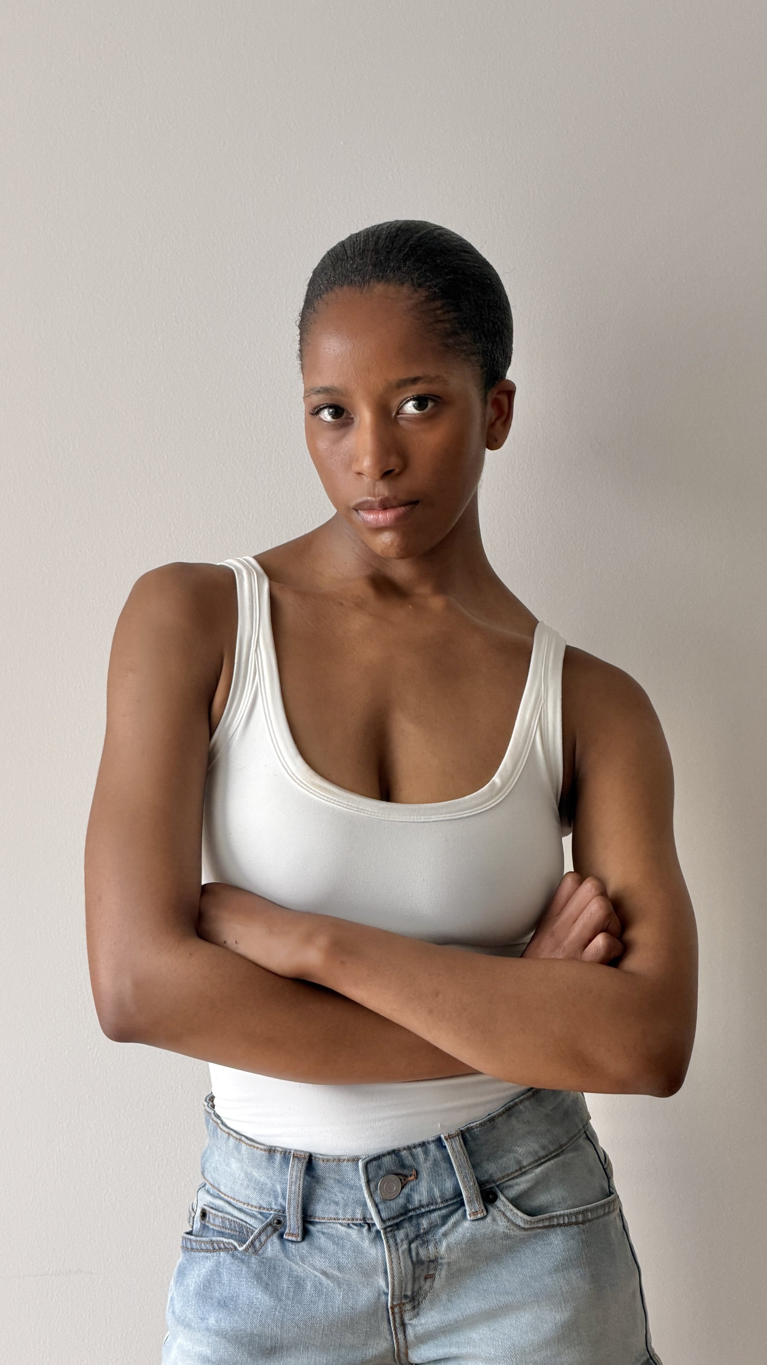 A young woman with dark hair pulled back, wearing a white tank top and light blue jeans, standing with her arms crossed against a plain white wall.