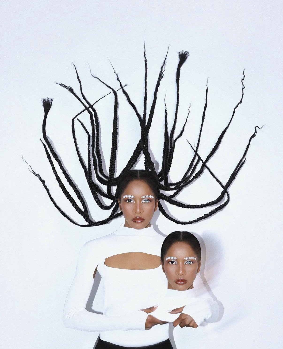 Two women wearing white outfits with cutouts, decorated with silver eye makeup, and posing against a white background. They have long braids spread out behind them.