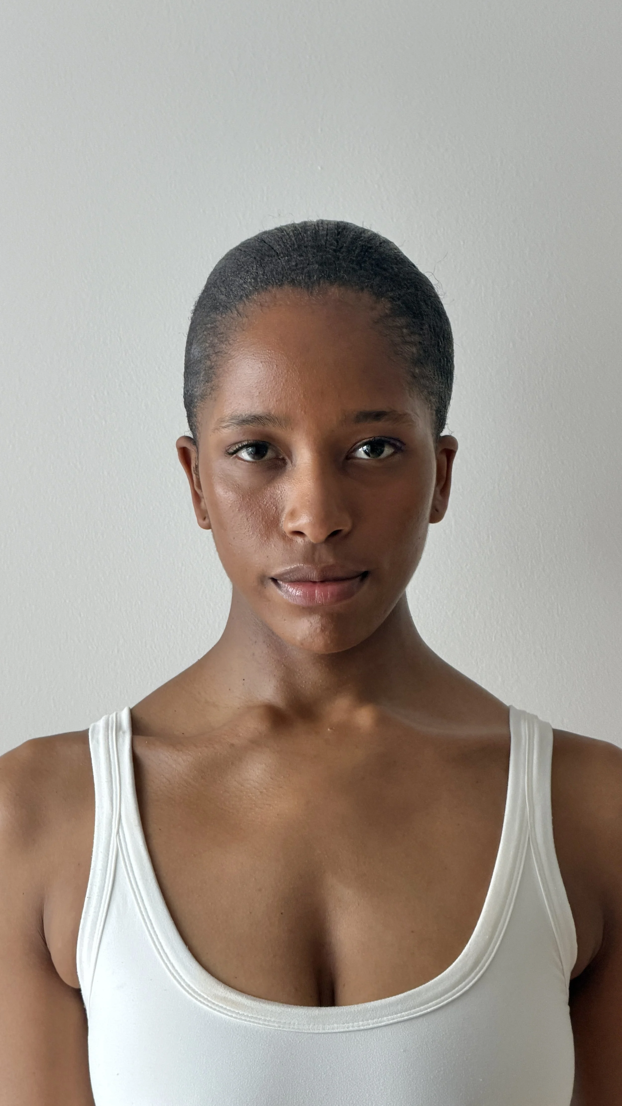 A woman with short natural hair, wearing a white tank top, standing against a plain light-colored wall.