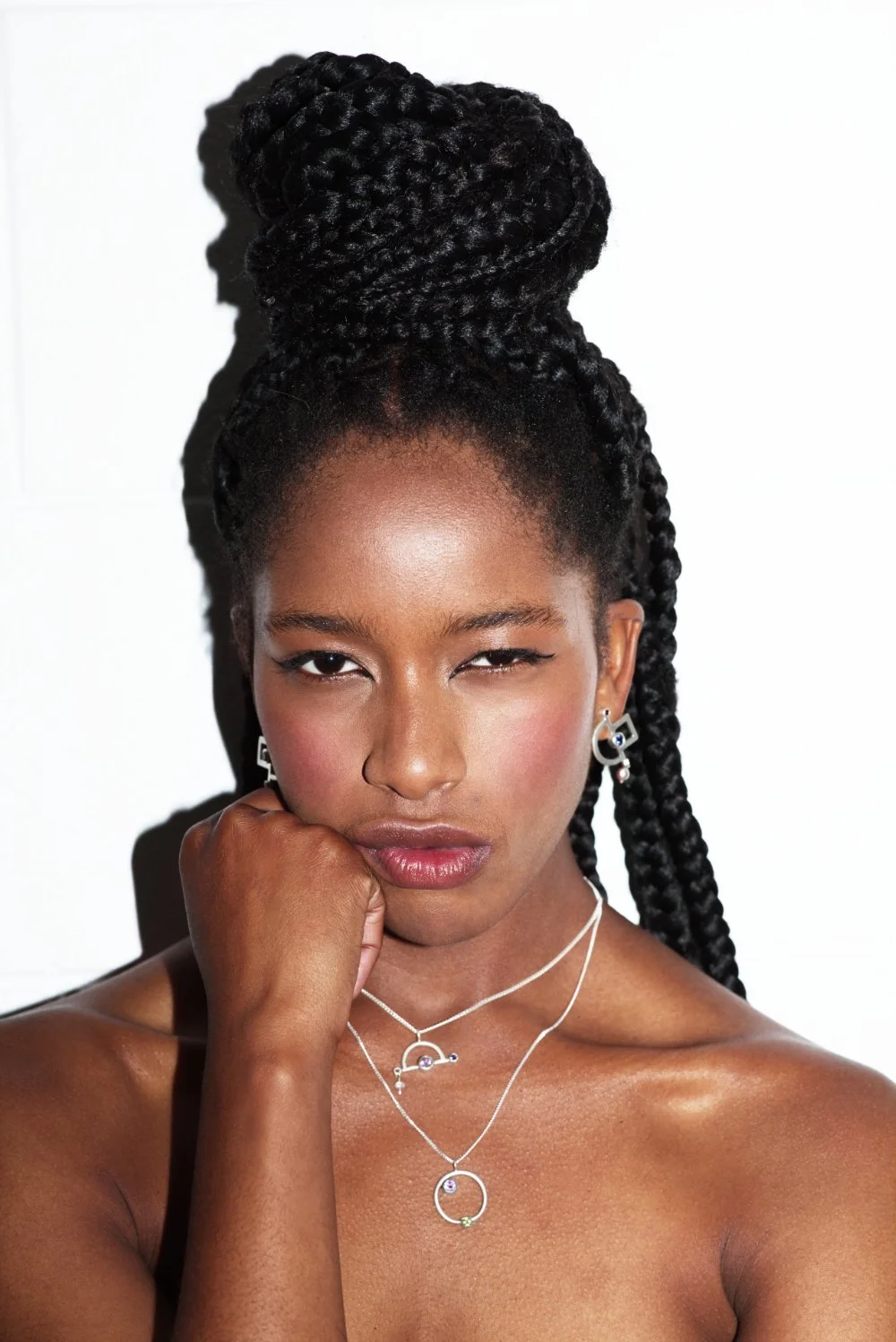 Close-up of a woman with dark skin wearing jewelry, with her hair styled in thick braided twists piled on top of her head. She has a confident expression with her chin resting on her hand, looking directly at the camera.