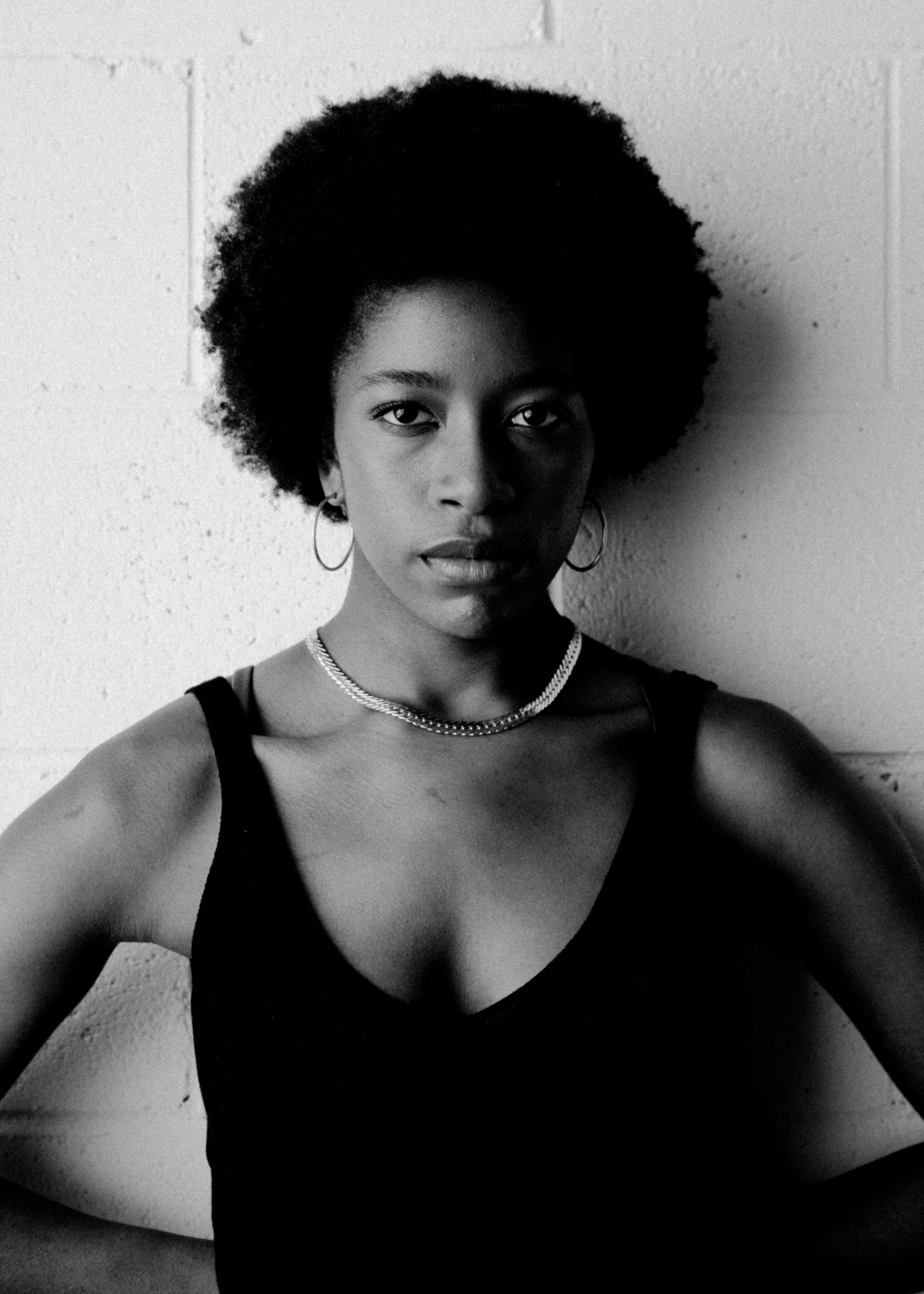 Black and white portrait of a woman with an afro hairstyle, wearing hoop earrings, a chain necklace, and a sleeveless top, looking directly at the camera against a textured wall background.