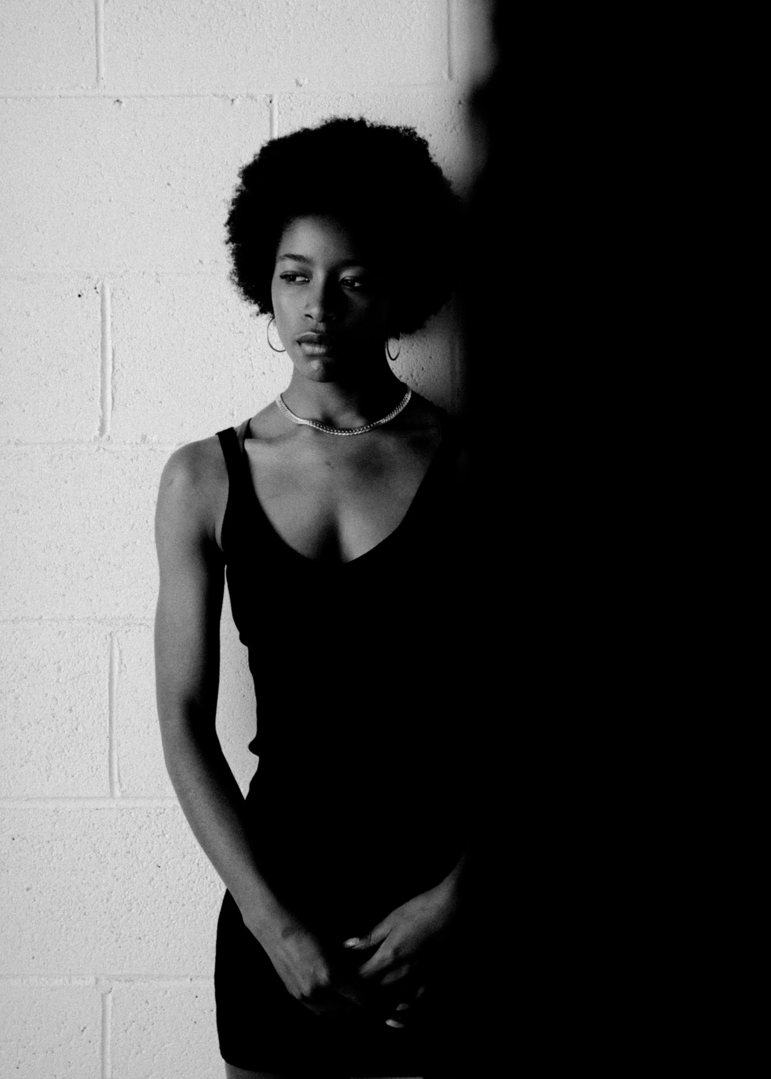 Black-and-white photo of a woman with an afro hairstyle standing against a plain wall, partially obscured by a shadow on the right side.