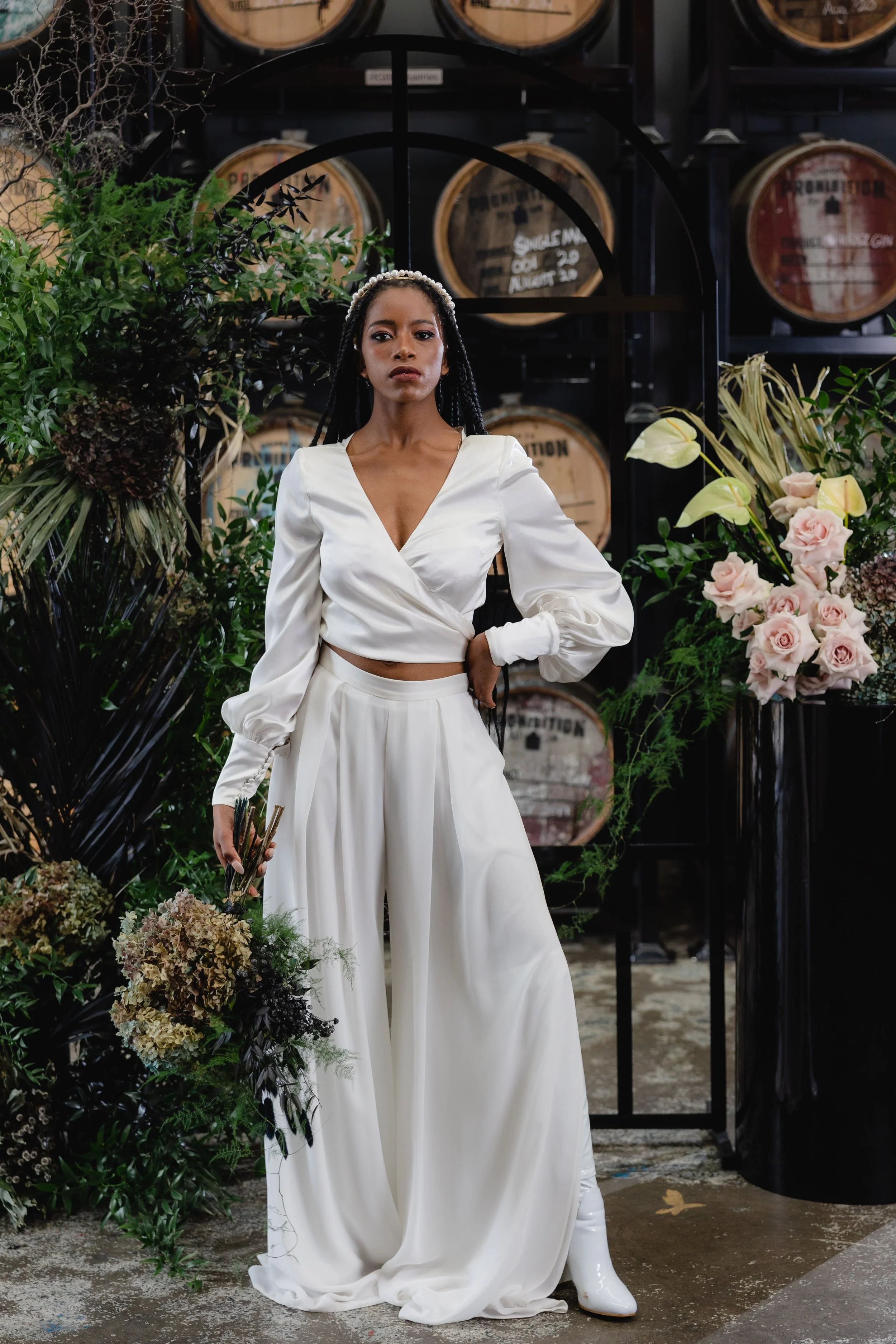 A woman with braids, wearing a white, wrap-style blouse and flowing white pants with a headband of pearls, standing among flowers and greenery in front of shelves with whiskey barrels.