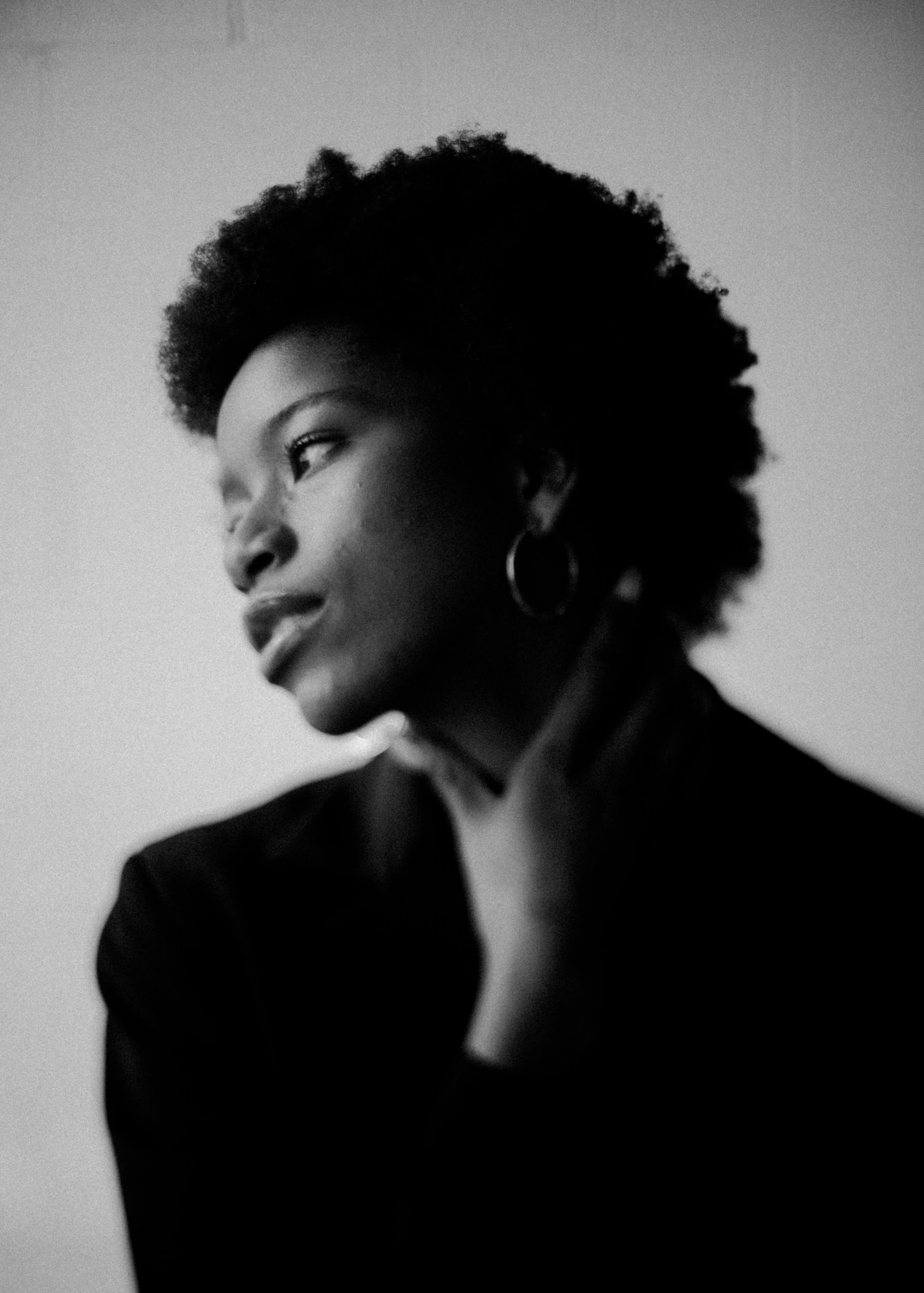 Black and white portrait of a woman with natural curly hair, wearing a dark blazer and hoop earrings, looking contemplative with her hand near her chin.