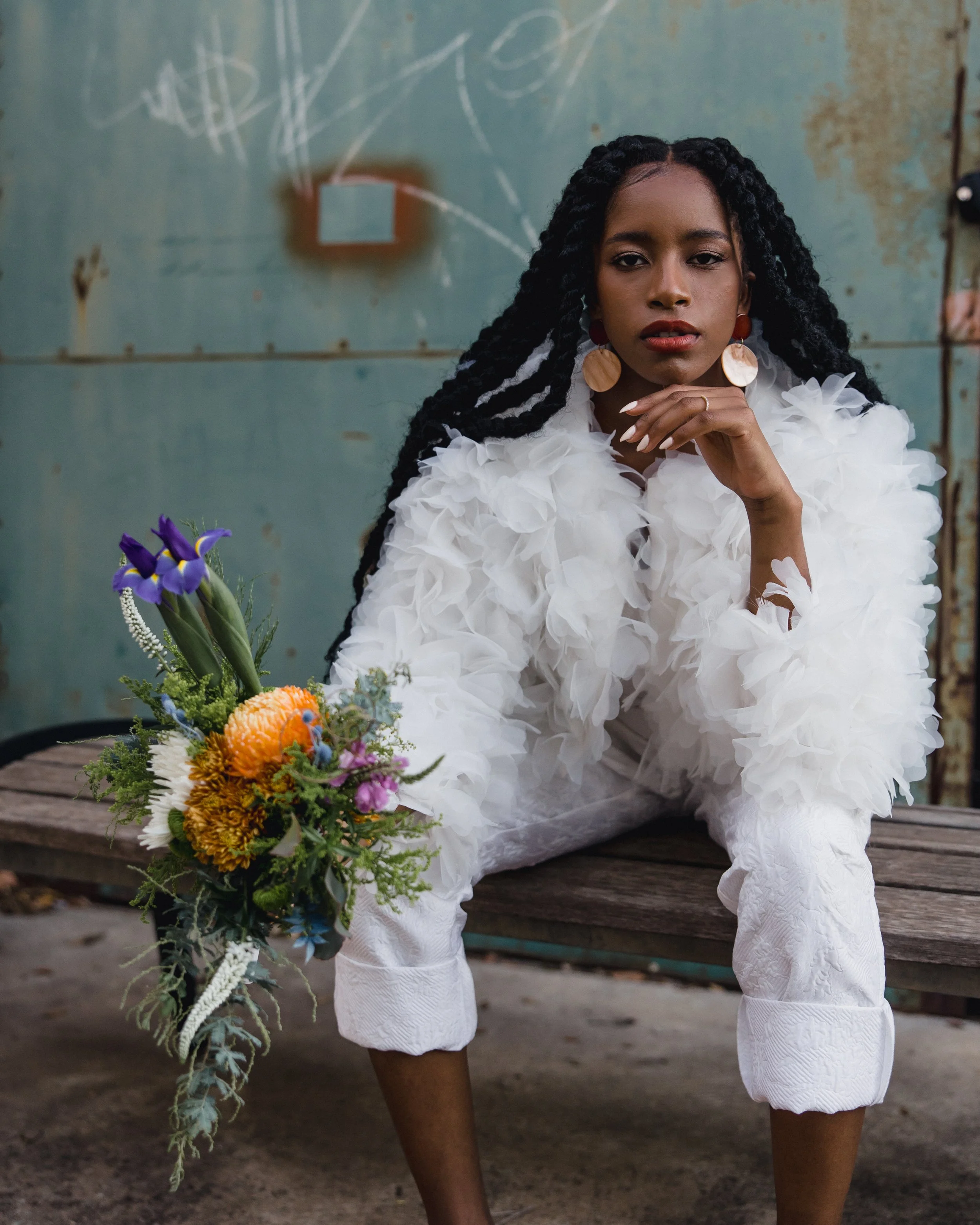 A woman sitting on a wooden surface in front of a weathered wall with chalk markings. She is dressed in a white, textured outfit with ruffled sleeves. She has long braided hair, large earrings, and is resting her chin on her hand. There is a colorful