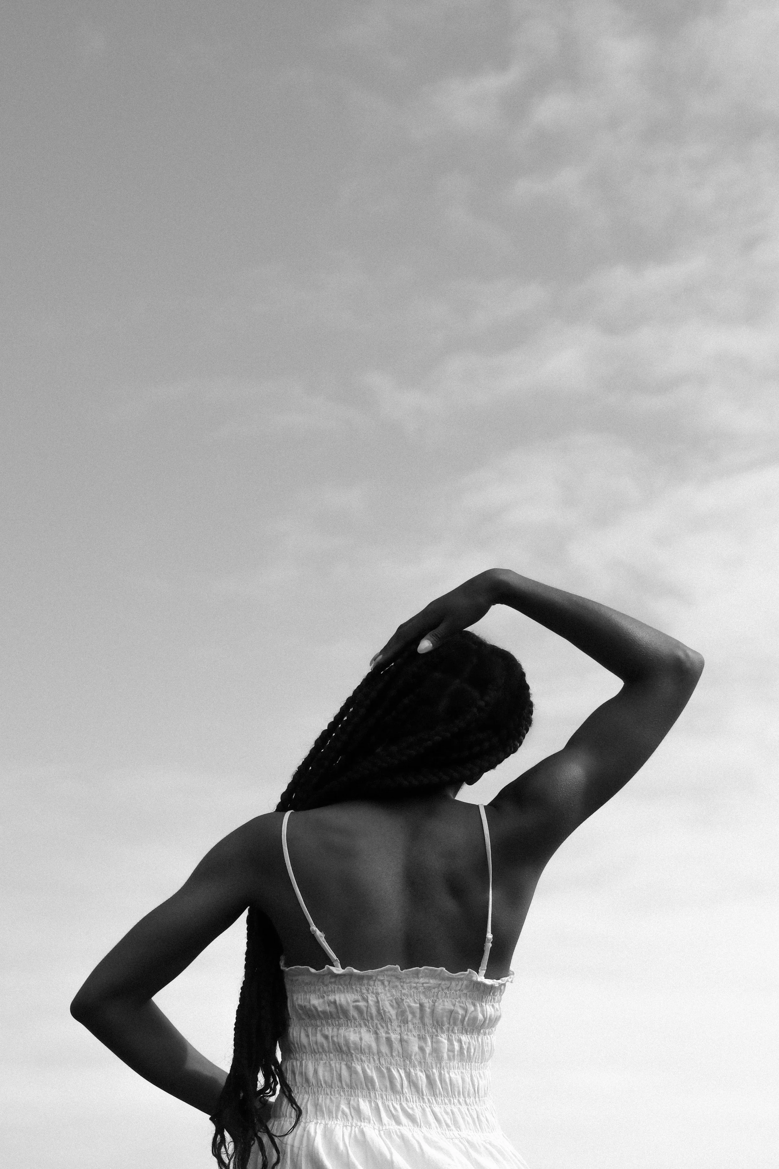 A woman with long braided hair is standing outdoors under a cloudy sky, wearing a sleeveless top and a loose, textured skirt, with one arm raised and her hand resting on her head.