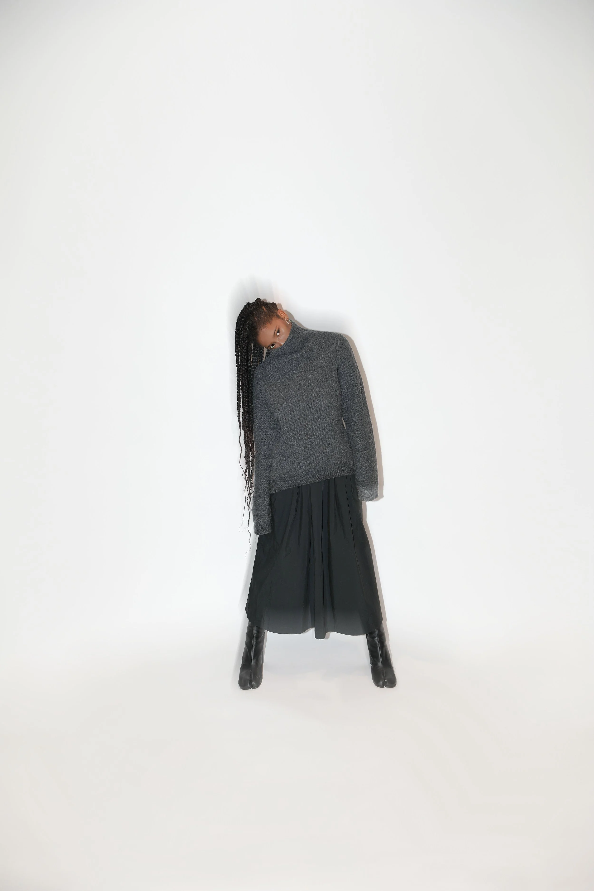 A woman with long braids wearing a grey turtleneck sweater, a black pleated skirt, and black high-heeled boots, standing against a plain white background with her head turned and one arm resting by her side.