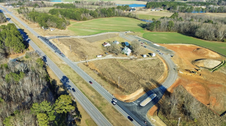 Aerial view of a rural area with roads, open fields, and some buildings surrounded by trees.