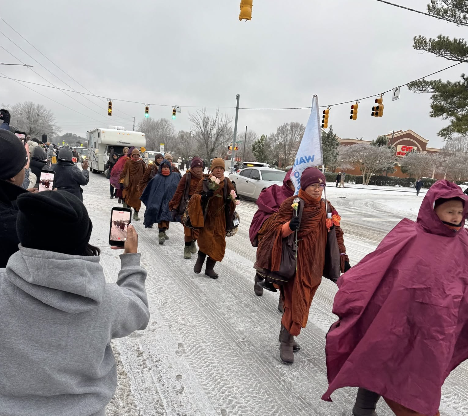 A walk of monks in brown robes carrying flowers during a parade on a snowy day, with spectators capturing the event on their phones. There are traffic lights and a CVS store in the background.