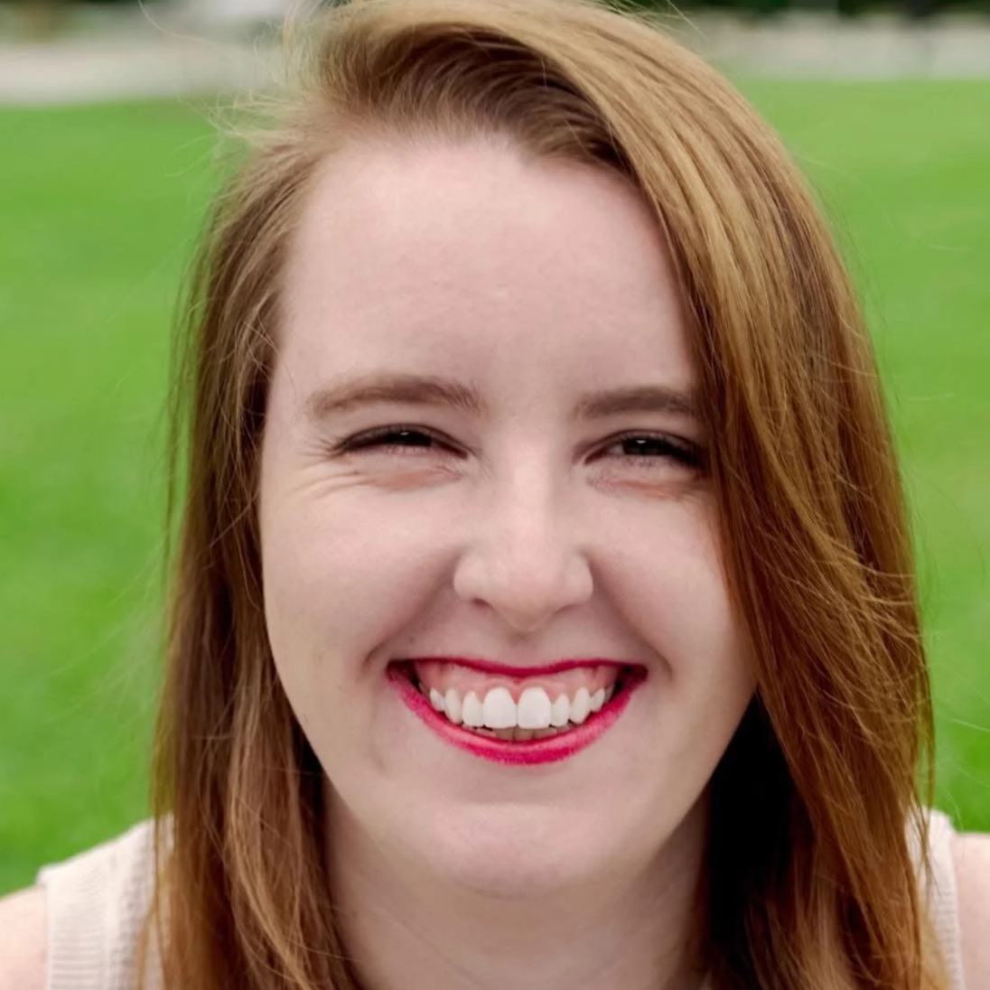 Close-up of a smiling woman with red hair and red lipstick, outdoors with green grass background.