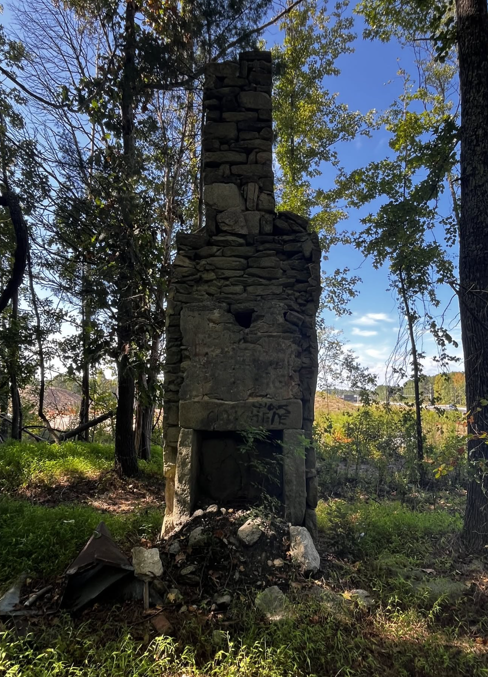 Old abandoned stone chimney in a wooded area with trees and blue sky in the background.