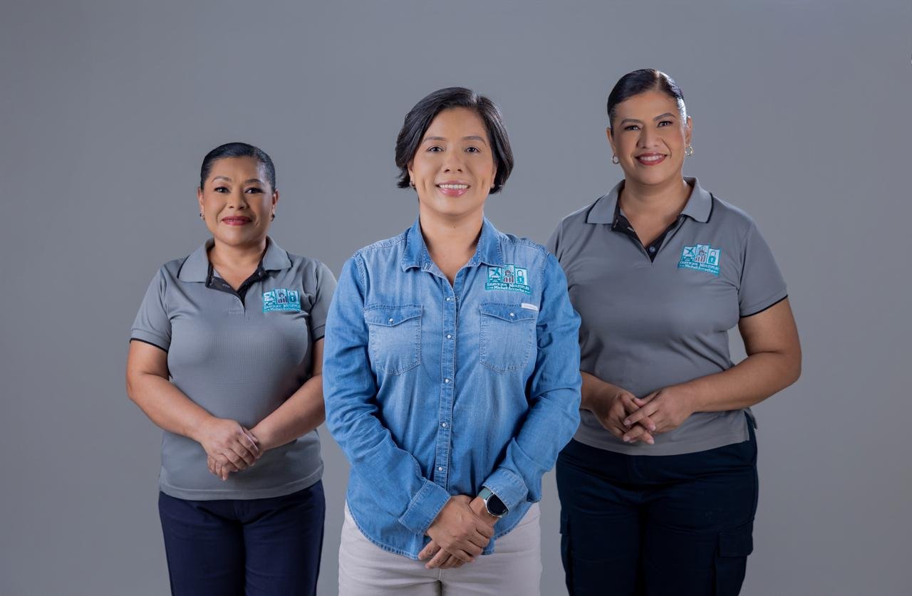 Tres mujeres de pie, sonrientes, con uniformes grises y una en manga larga azul, en un fondo gris liso.