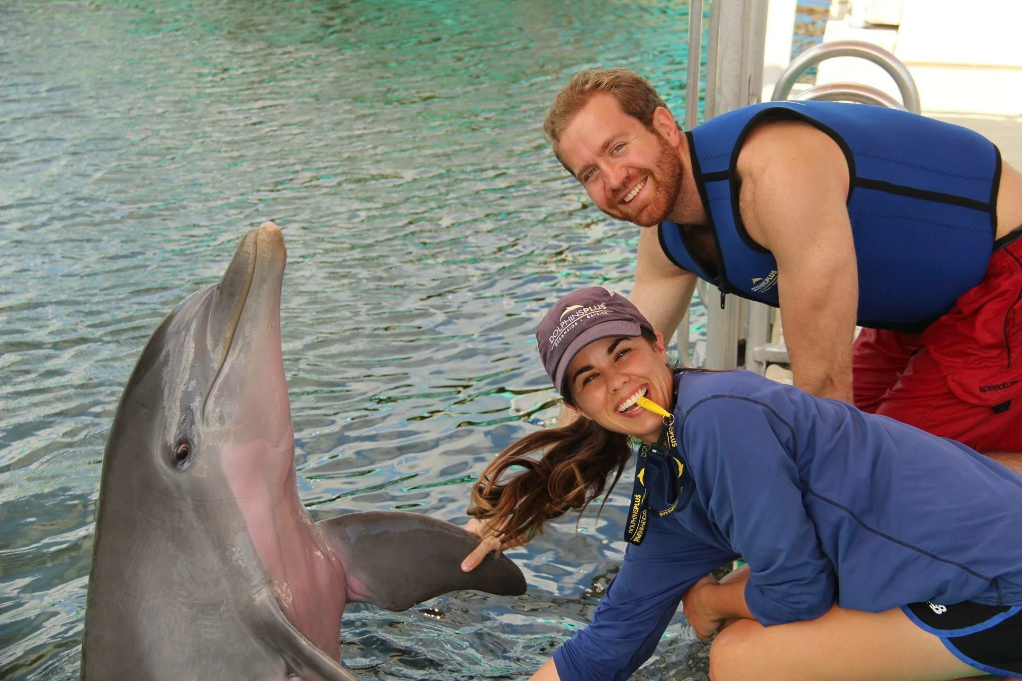 Two smiling young adults smiling with a dolphin in a body of water.