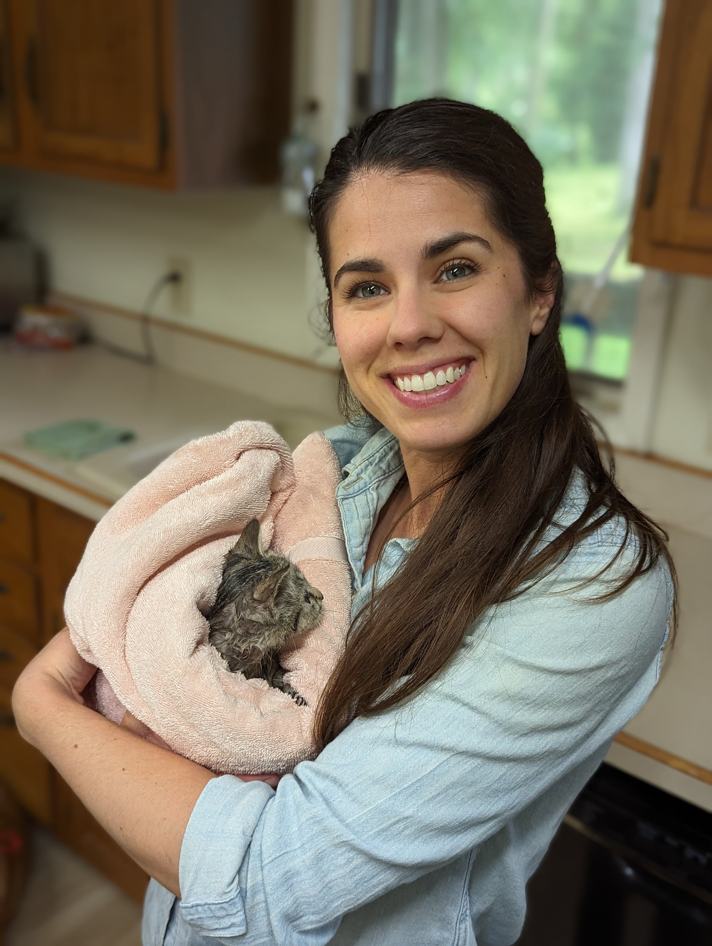 A woman with long brown hair smiling and holding a small kitten wrapped in a pink towel inside a room with wooden cabinets and a window with a view of greenery.