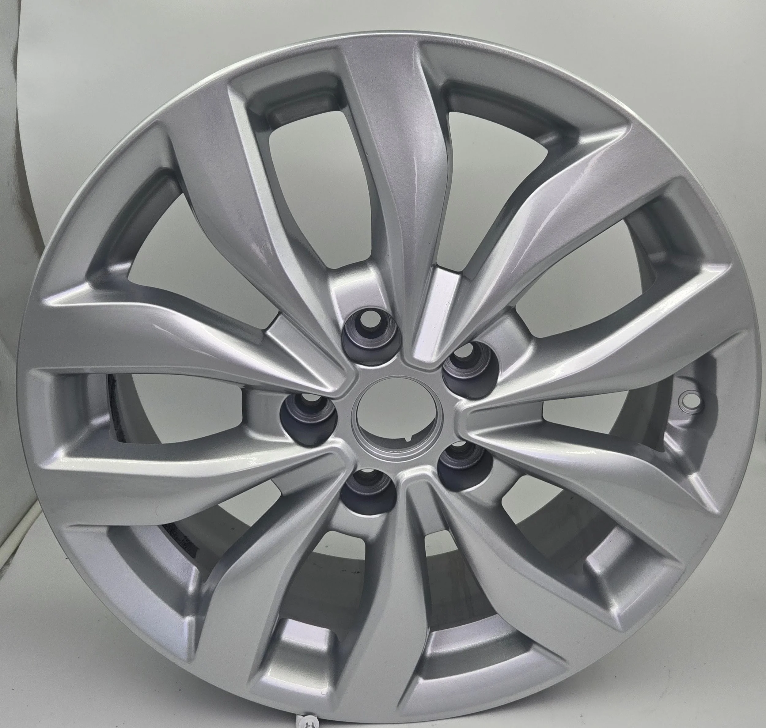 Silver car wheel rim with multiple spokes and a central hole, placed on a gray surface and leaning against a flat background.