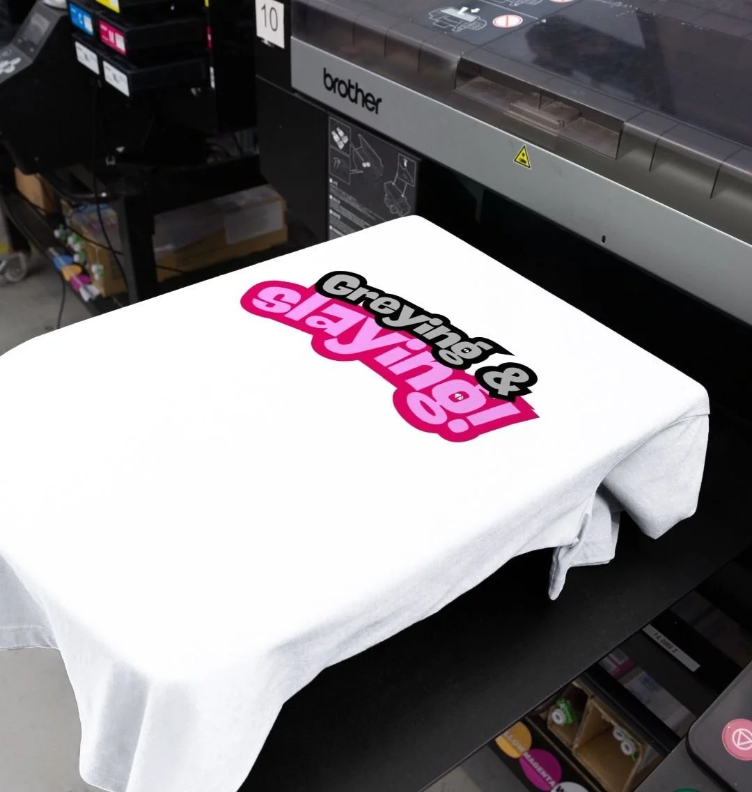 A white T-shirt being printed with the text 'Grevinet & RayJin' in black and pink, lying flat on a printer or heat press machine.