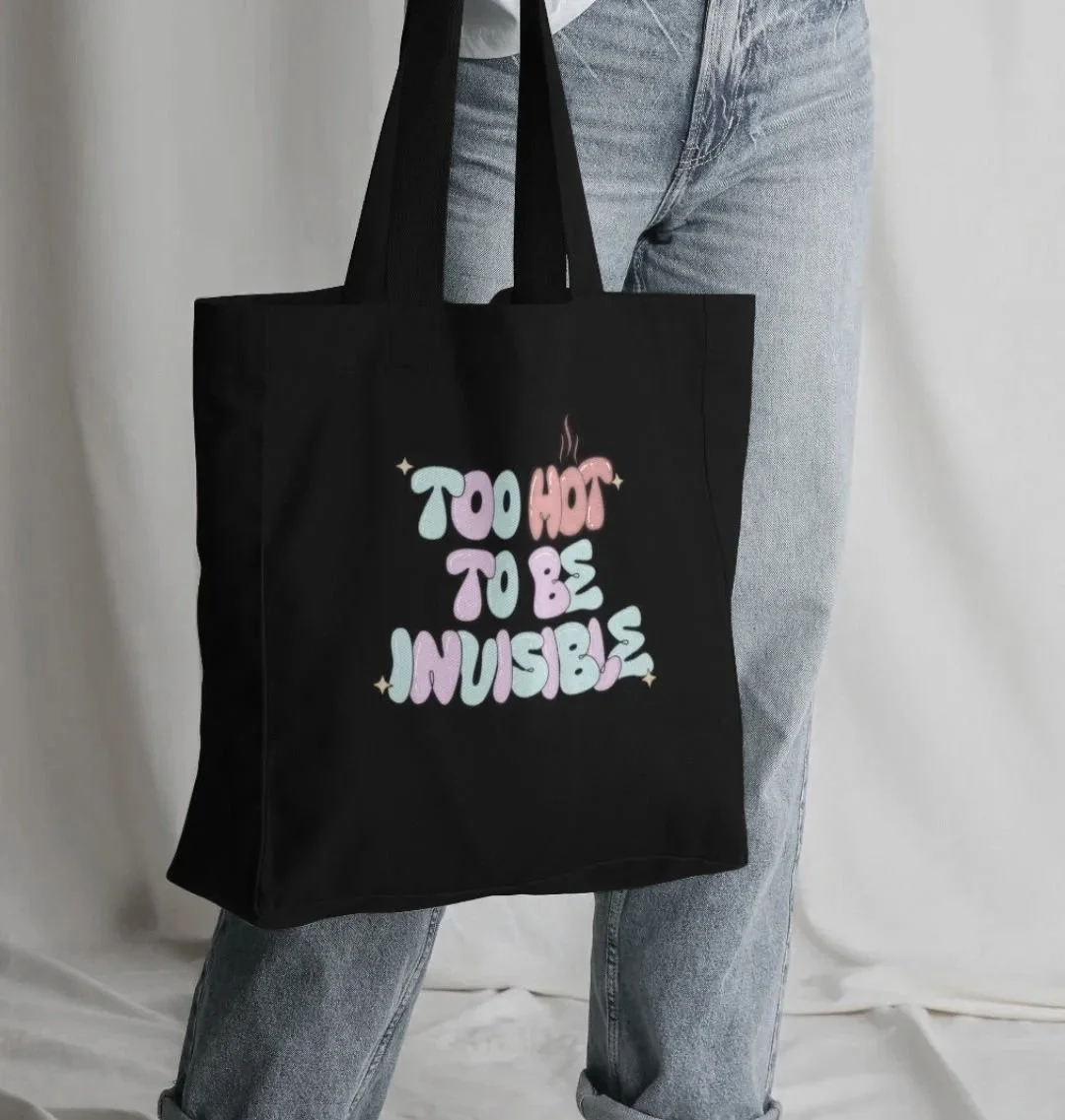 Person holding a black tote bag with colorful text that reads 'Too Hot To Be Invisible'.