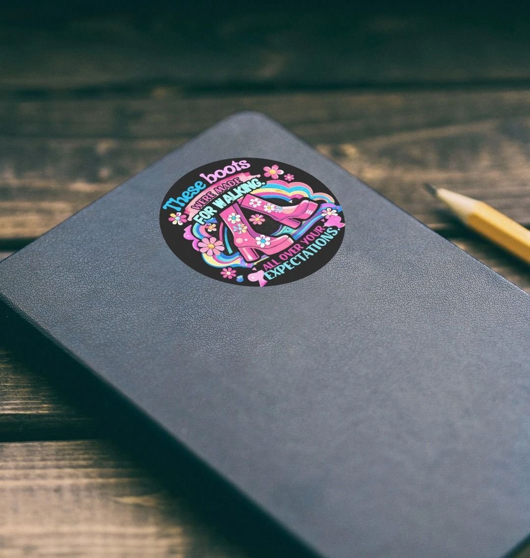 A gray notebook with a colorful sticker featuring pink high heels, flowers, and a rainbow, with the text 'These boots were made for walking, all over your expectations'.