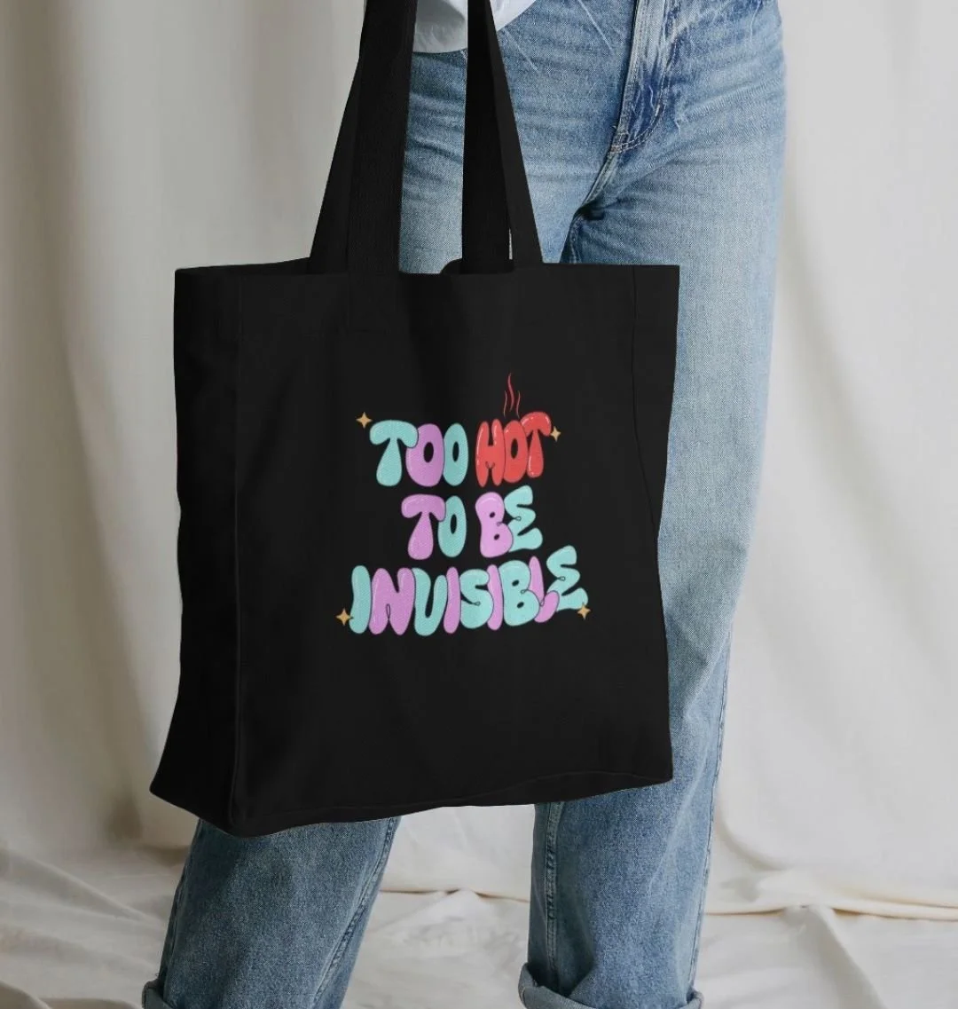 Person holding a black tote bag with colorful text that reads 'Too Hot to be Invisible' against a plain background.