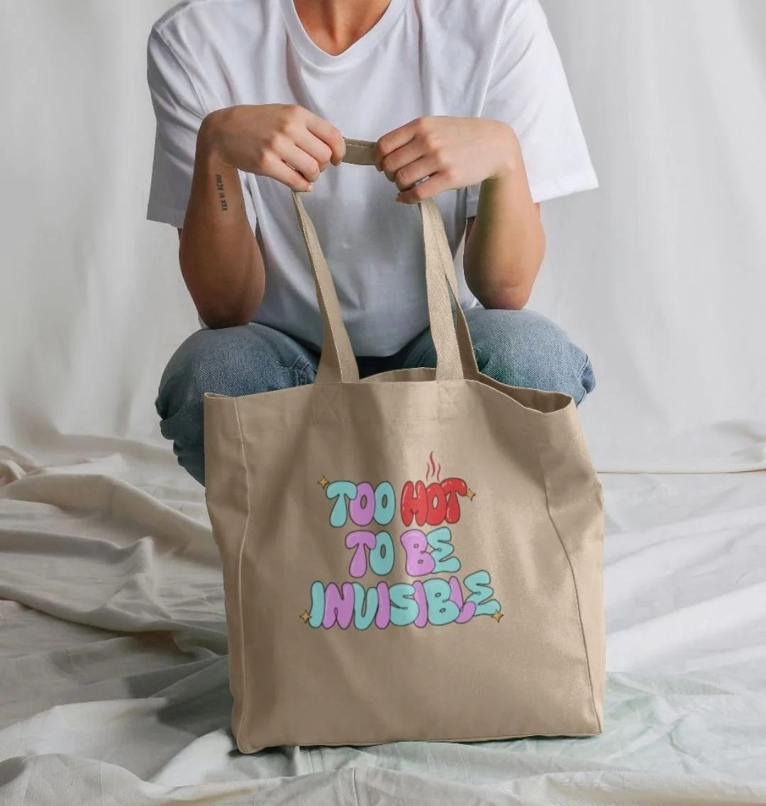 A person sitting on a bed holding a beige tote bag with colorful text that says "Too Hot to Be Invisible" and steam above the word "Hot."