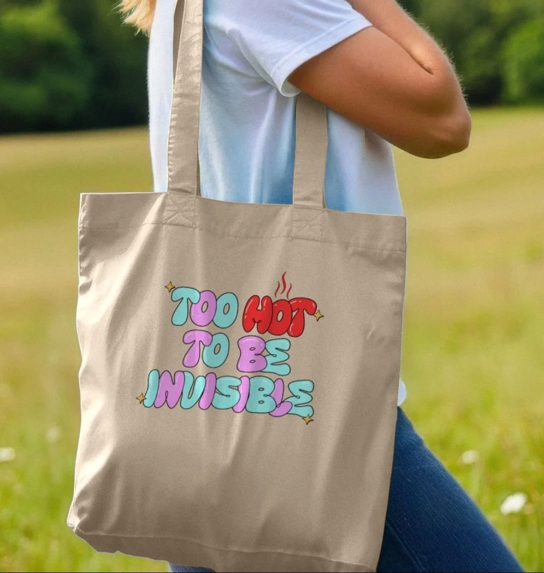 A person wearing a white t-shirt and blue jeans carrying a beige tote bag with the phrase 'TOO HOT TO BE HUMBLE' in colorful bubble letters, standing outdoors in a grassy area.