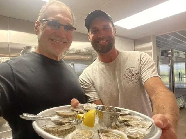 Two smiling men in a kitchen holding a plate of oysters with lemon wedges, one wearing glasses and a black shirt, the other with a cap and a gray T-shirt.