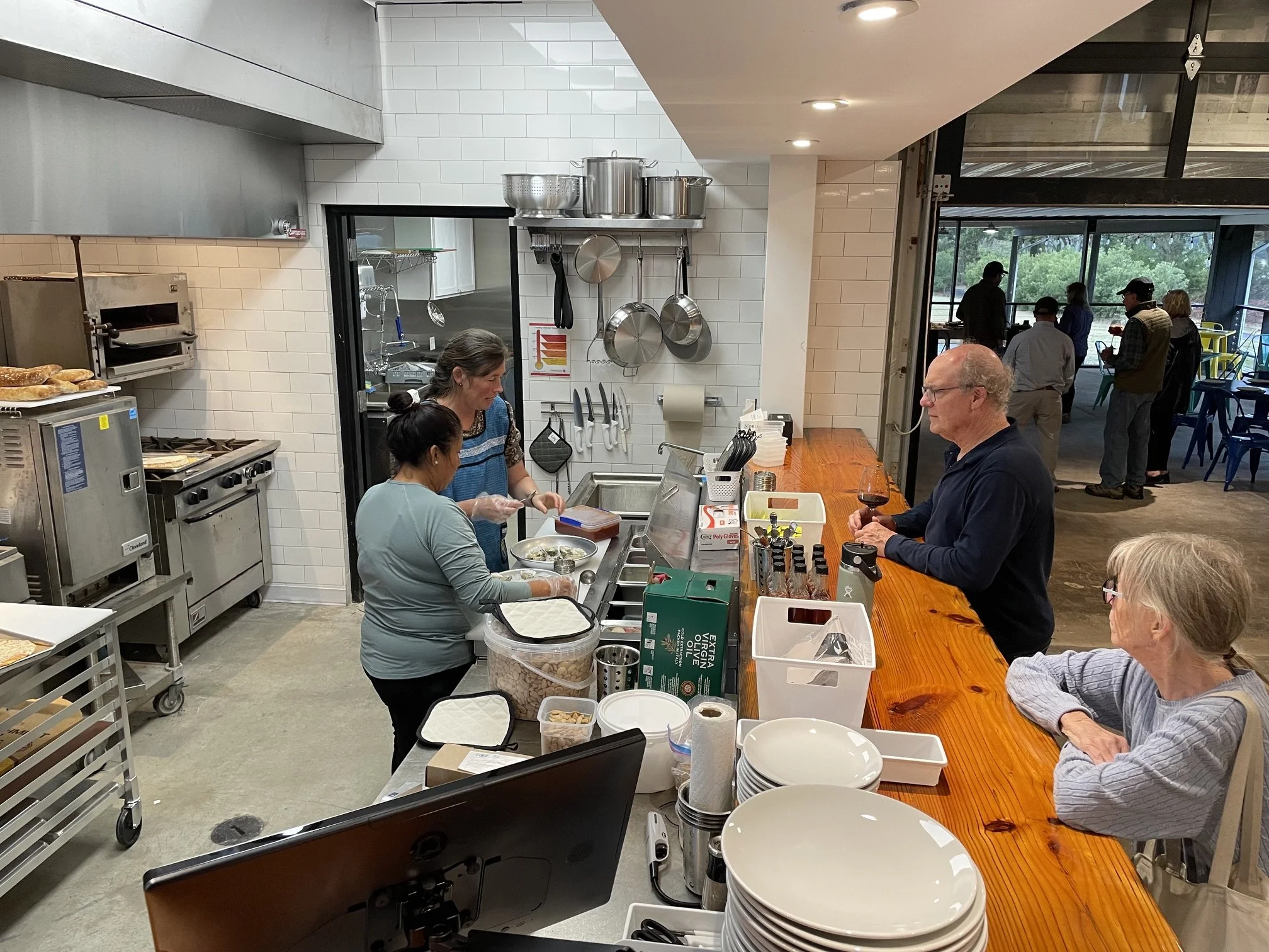 People serving food and drinking at a restaurant counter while others are seated and socializing in the background.