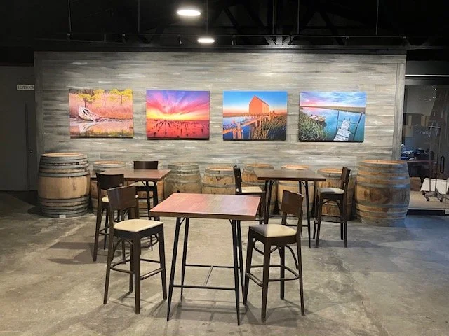 Interior of a modern room with a gray wooden accent wall, decorated with five landscape or abstract paintings. There are wooden barrel tables and black chairs arranged around a pink table, with additional barrels in the background.