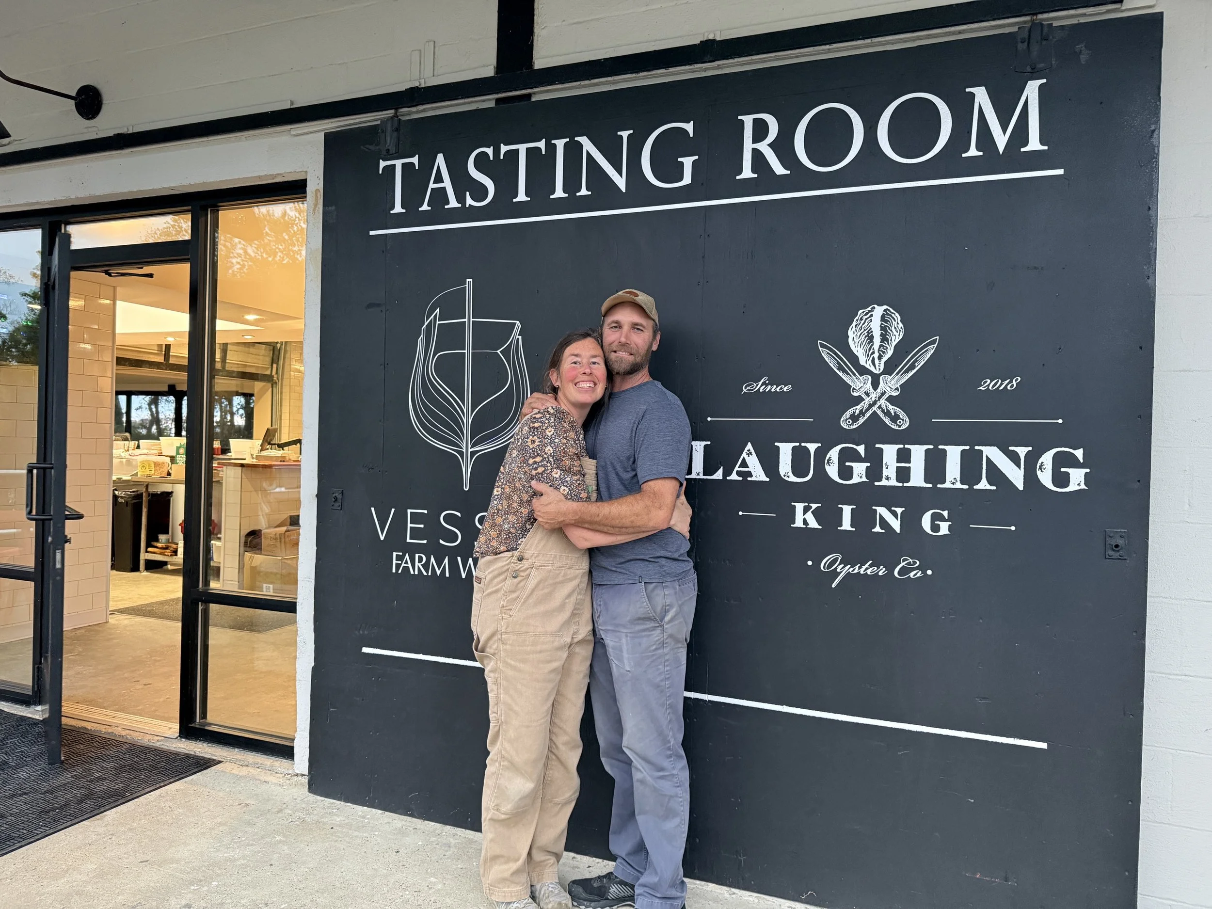 A man and a woman embracing and smiling outside a building with a large sign that reads "Tasting Room". The building has an open door revealing the interior with tables and some items on the counter.