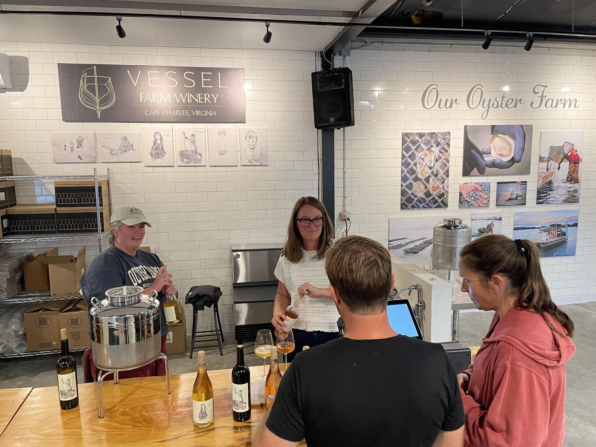 People attending a wine tasting event at Vessel Farm Winery in Cape Charles, Virginia. Several bottles of wine and wine glasses are on the table, with a white brick wall and artwork in the background.