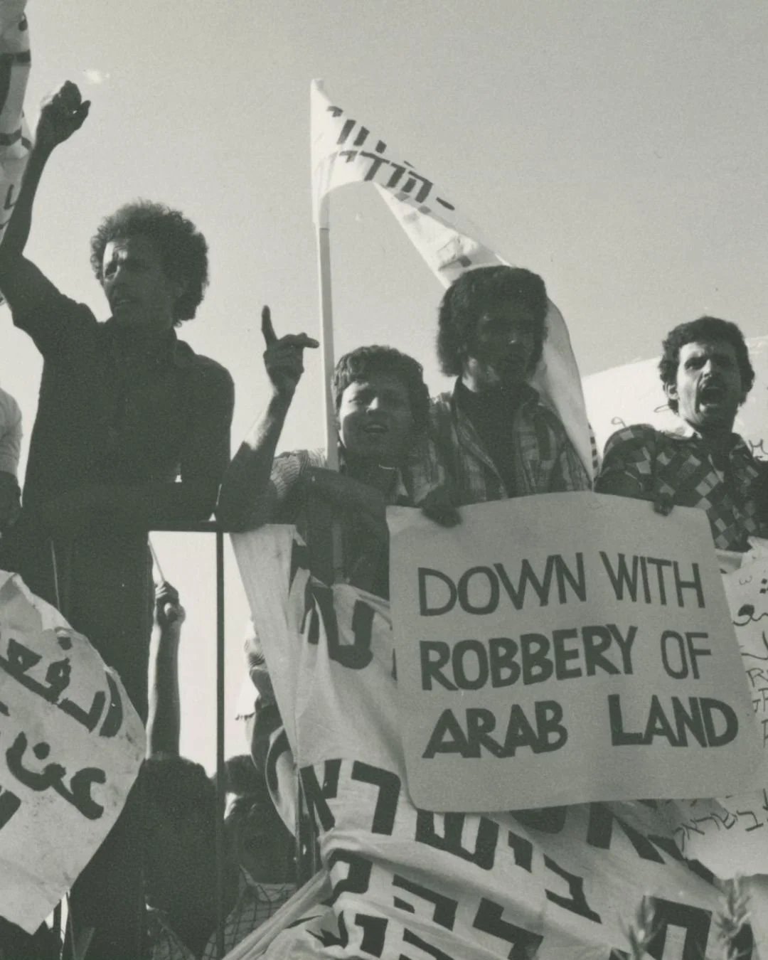 A black and white photograph of protesters holding signs and banners, one sign reads 'Down with robbery of Arab land', with some individuals making gestures or raising their fists in protest.