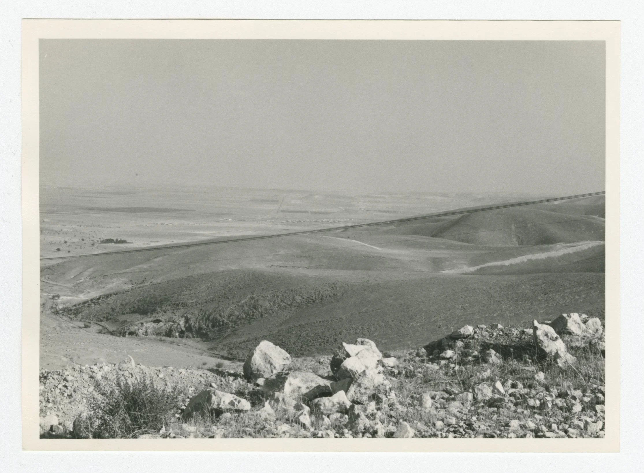 A black and white photograph of a desert landscape on a mountain road, with scattered rocks and sparse vegetation in the foreground and distant mountains in the background.