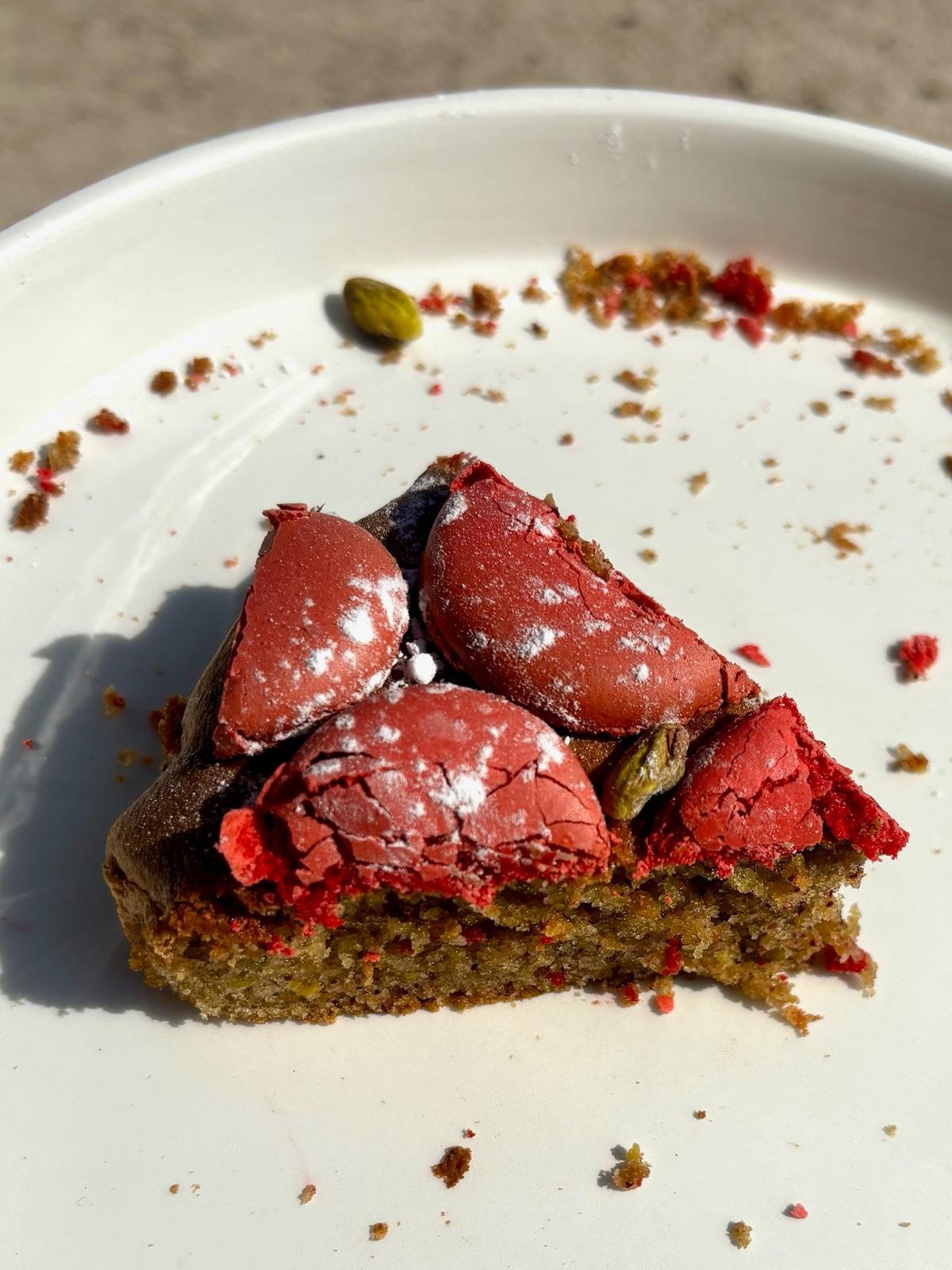 Part de tarte avec une croûte croustillante, garnie de pistaches et de morceaux de chocolat rouge, avec quelques miettes de gâteau sur une assiette blanche.