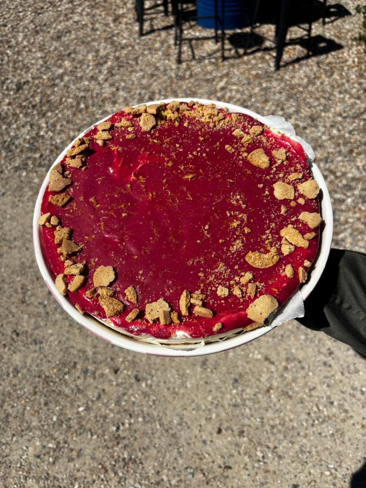 Gâteau à la crème de framboise avec des miettes de biscuits sur le dessus, tenu par une personne contre un fond de sol en gravier.