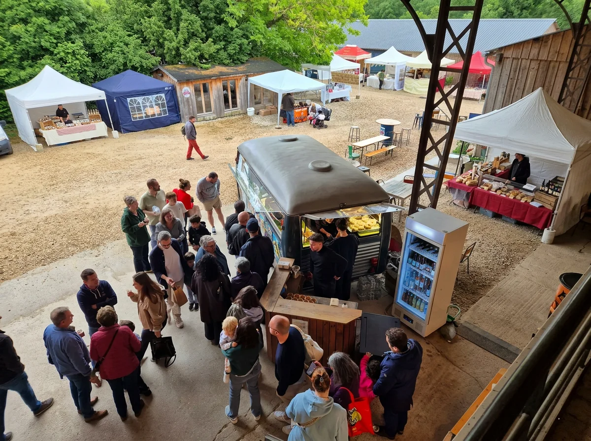 Marché en plein air avec plusieurs stands sous tentes colorées, une grande foule de personnes faisant la queue devant un camion food-truck avec de la nourriture en vitrine, et des stands avec produits locaux, le tout dans un environnement vert et boisé.