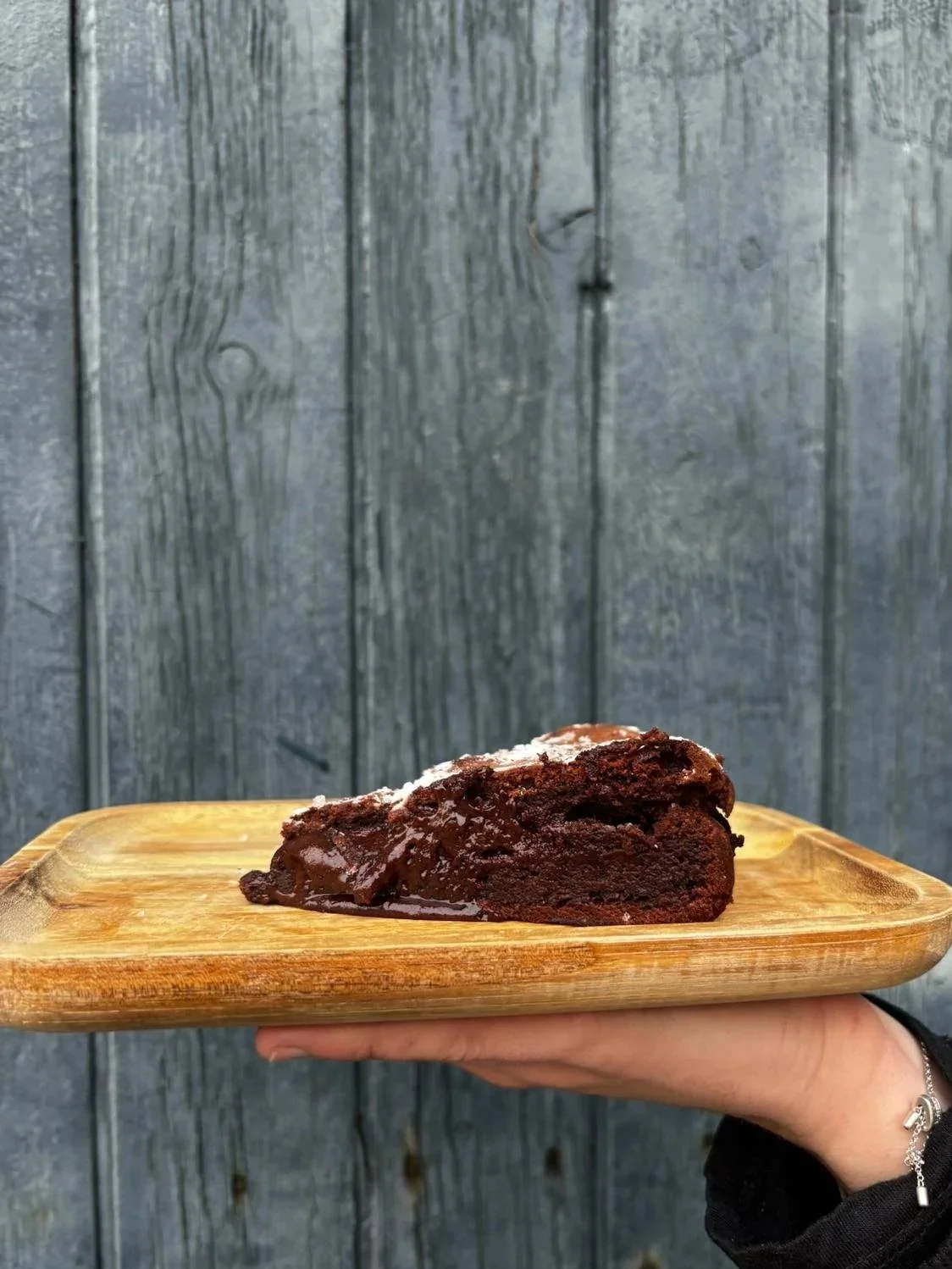 Part de gâteau au chocolat sur une planche en bois, tenue par une main contre un fond en bois.