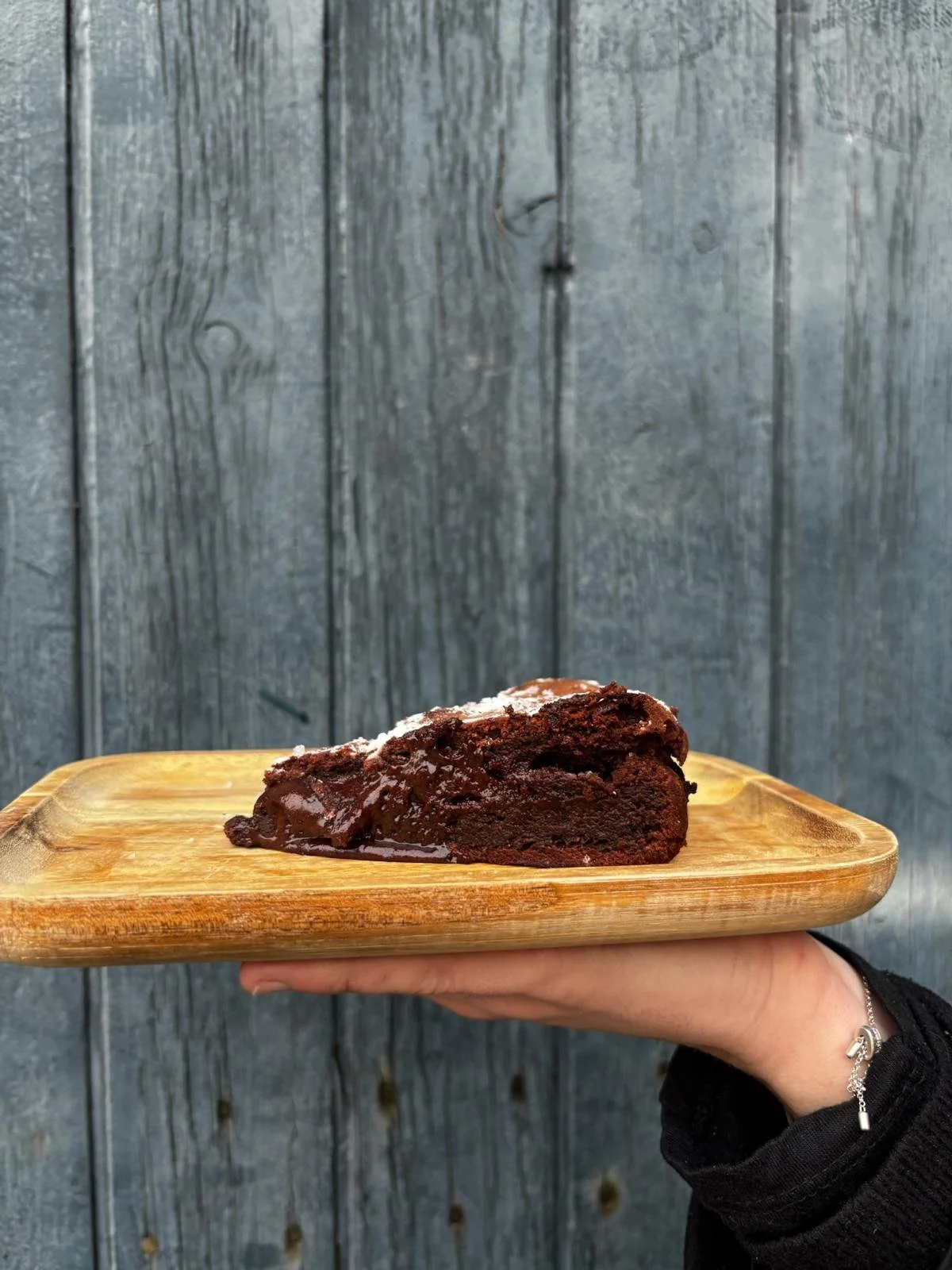 Une tranche de gâteau au chocolat sur une planche en bois, tenue par une main devant un fond en bois.