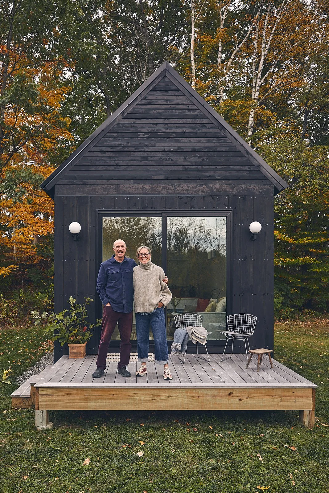 Two smiling plum prefab co-founders, standing on a small wooden deck outside an elegant black cabin with large glass doors, surrounded by autumn trees.