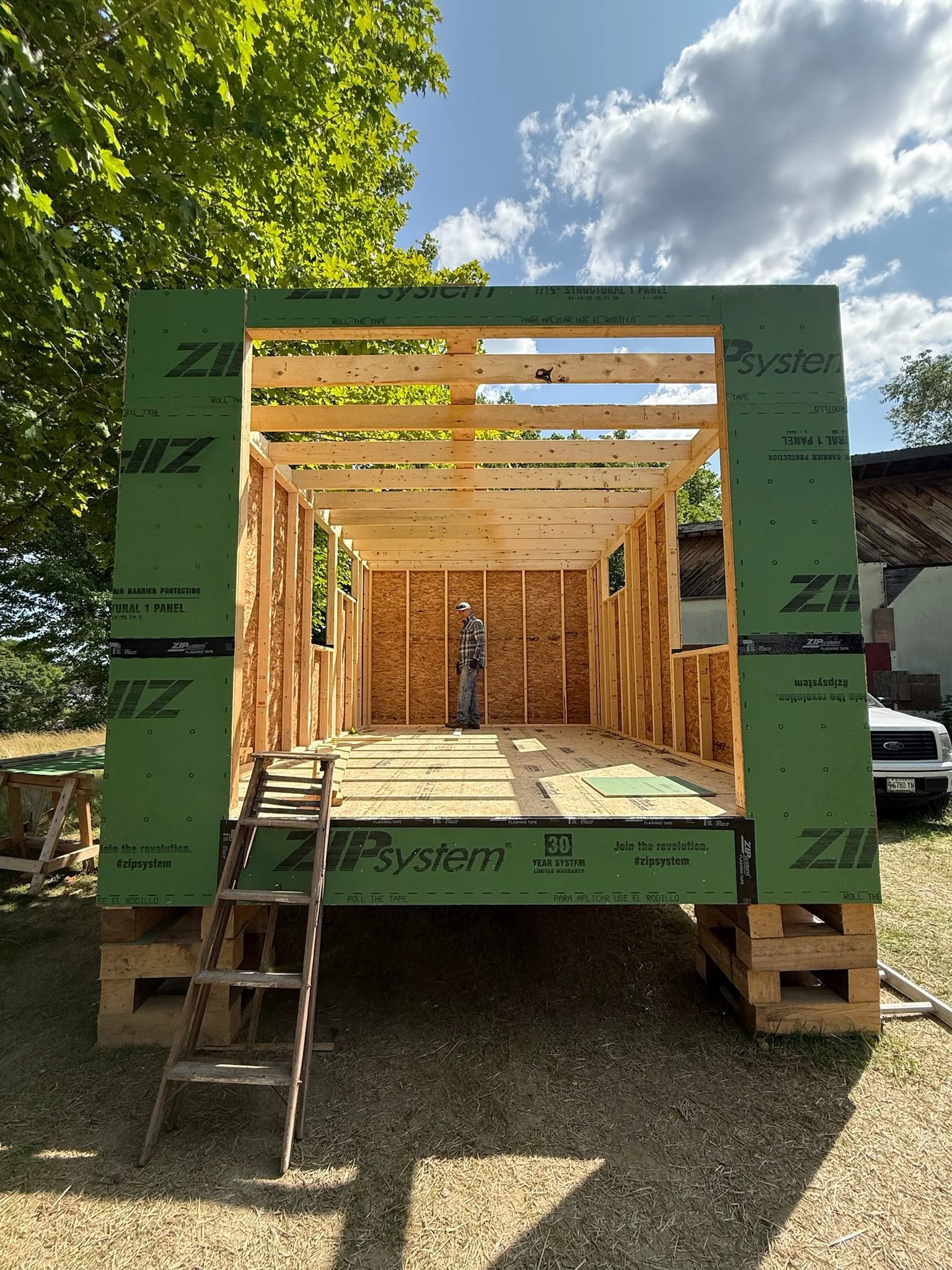 A wooden framing structure under construction, with a man standing inside, against a backdrop of trees and a partly cloudy sky.