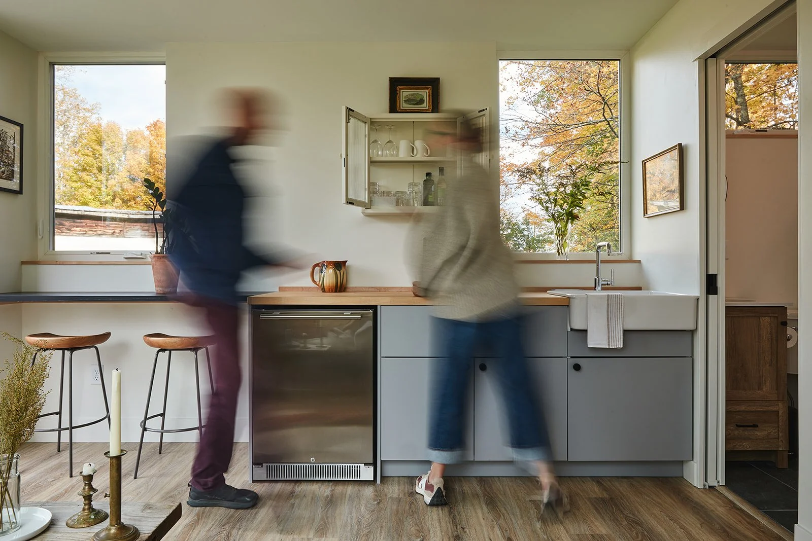 Blurry image of two people in a modern kitchen with large windows showing fall trees outside. One person is standing at the counter, while the other is walking past.