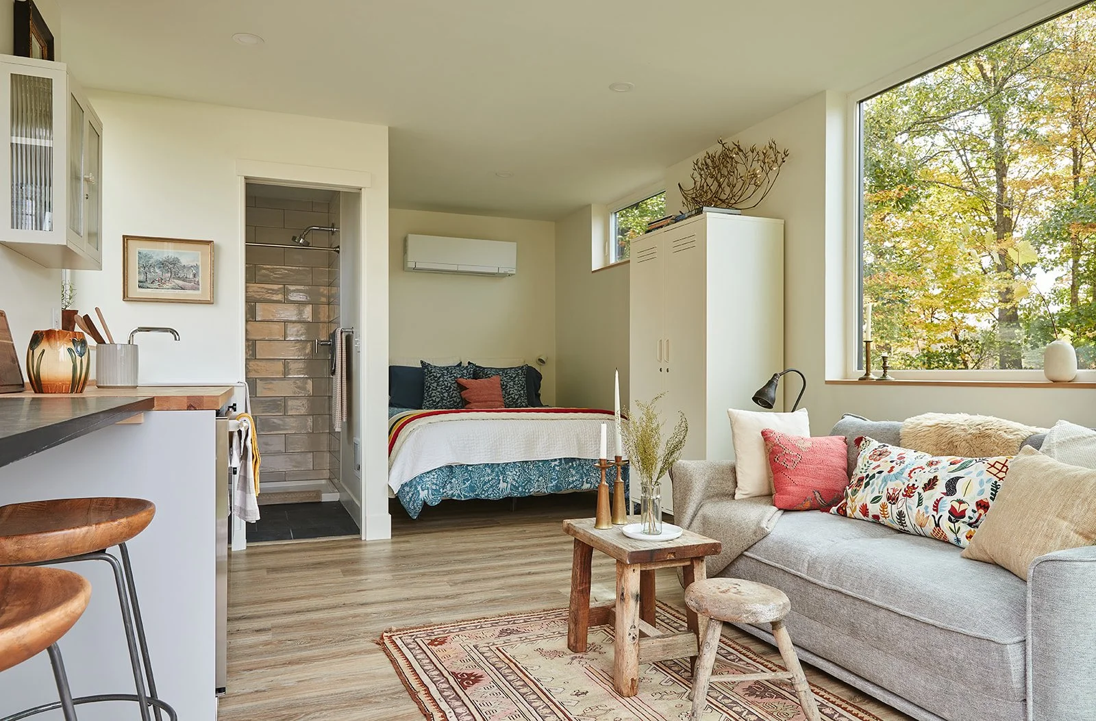 Open-concept living space with a gray sofa, colorful throw pillows, a small wooden coffee table with decorative candles and plant, large windows showing trees outside, a bed with throw pillows and a blanket, and some kitchen elements on the left.