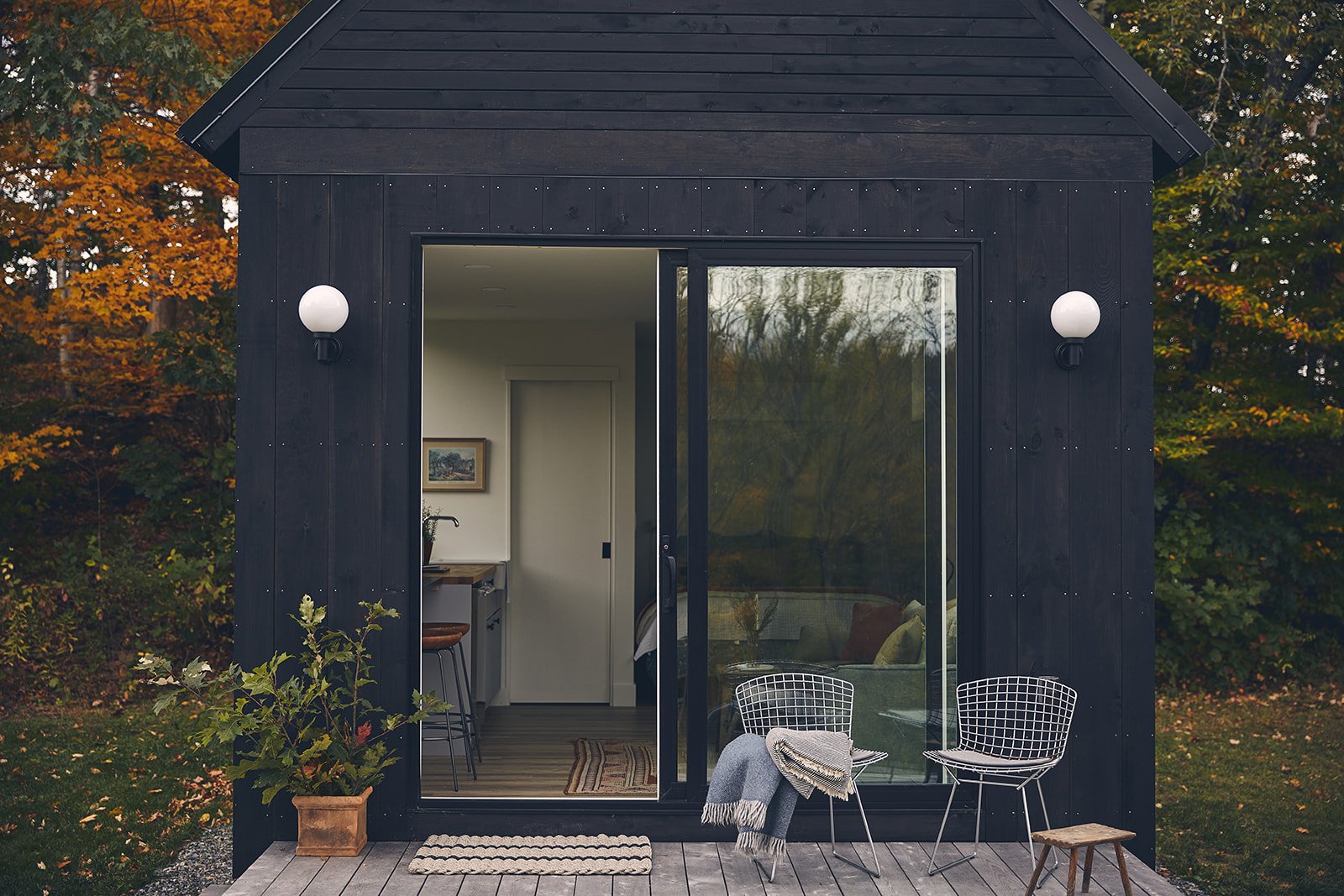 Small black modern wooden house with interior visible through glass sliding door, outdoor seating area with two wire chairs, a small wooden stool, a blanket, and a potted plant, surrounded by fall foliage.