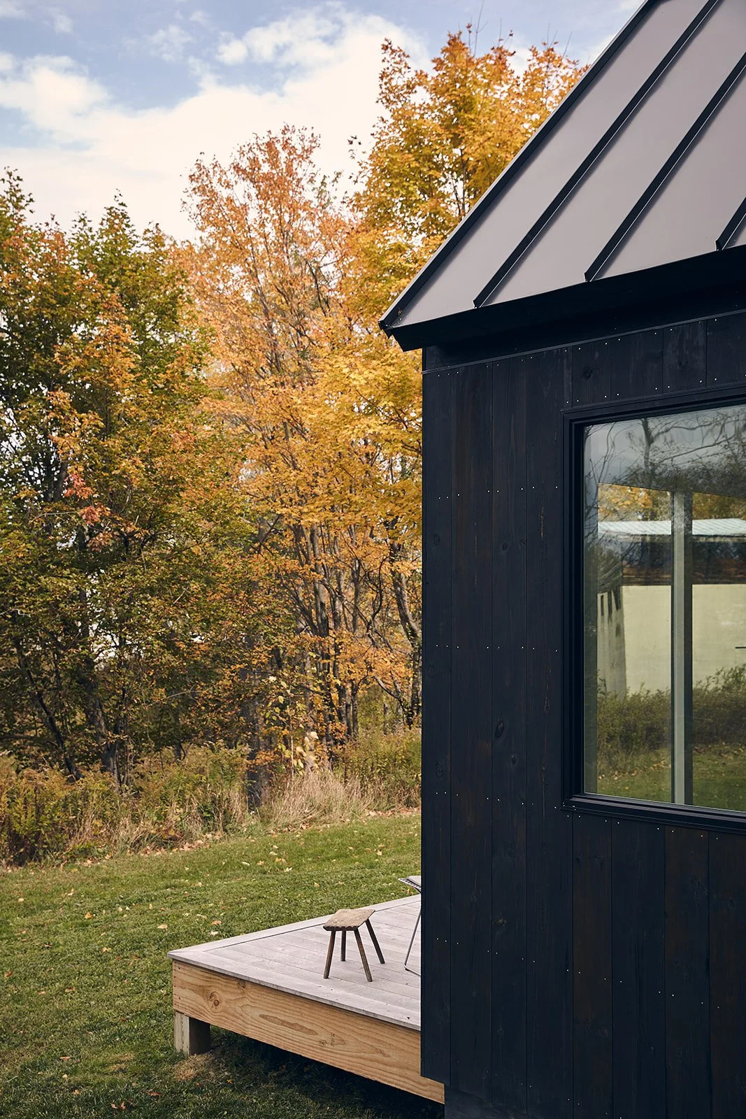 Side view of a modern black cabin with a large window, wooden deck, and a small stool outside, surrounded by trees with autumn foliage.