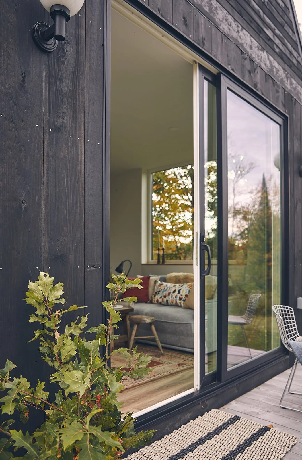 A view of a modern house with a sliding glass door open to a cozy interior living room. The house exterior features black wooden siding, a black wall-mounted light fixture, and greenery in the foreground. Inside, there is a grey couch with decorative
